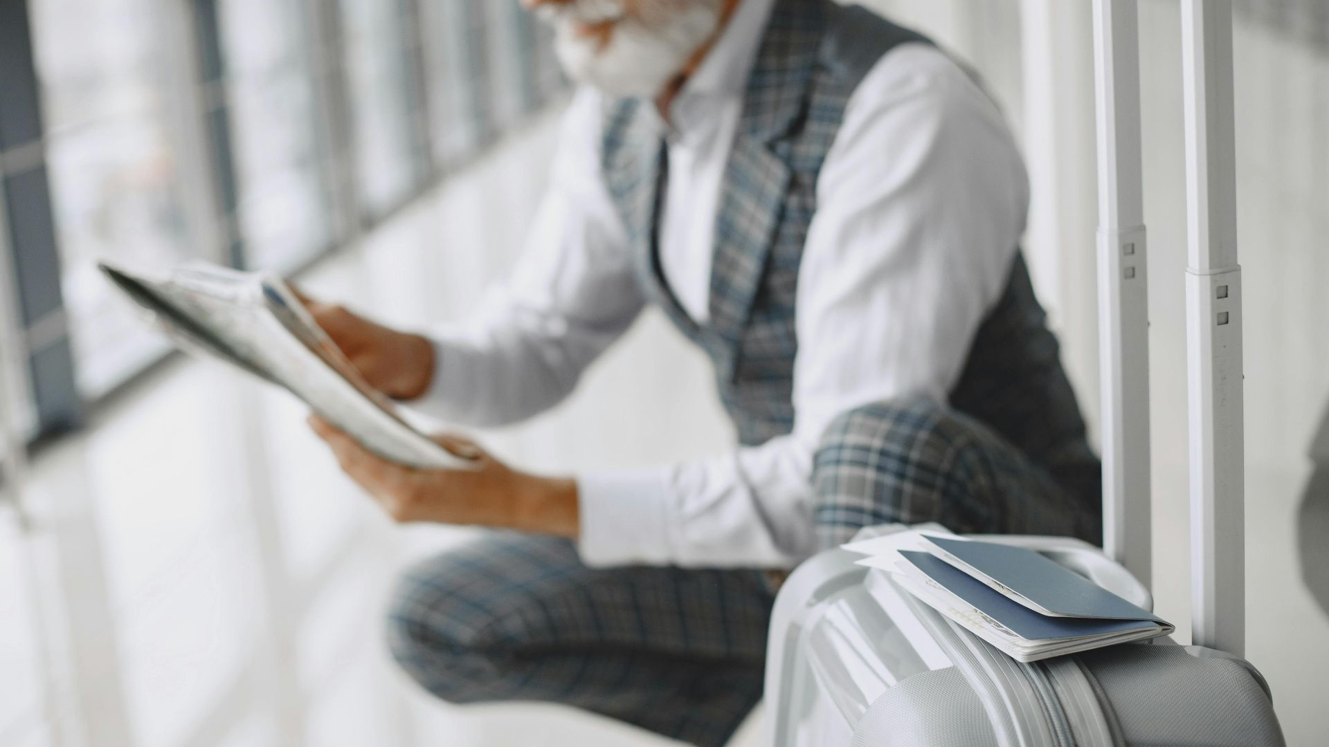 Adult man in plaid suit reads newspaper with suitcase at airport terminal.