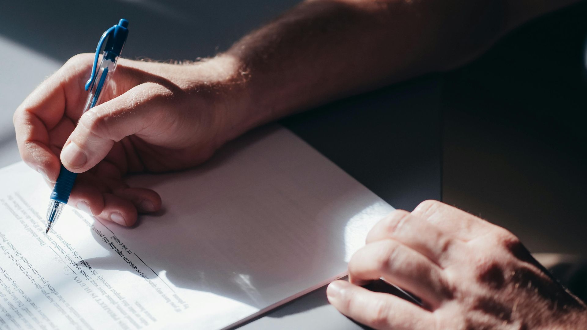 A close-up image of a person's hand signing a document with a blue pen in natural lighting.