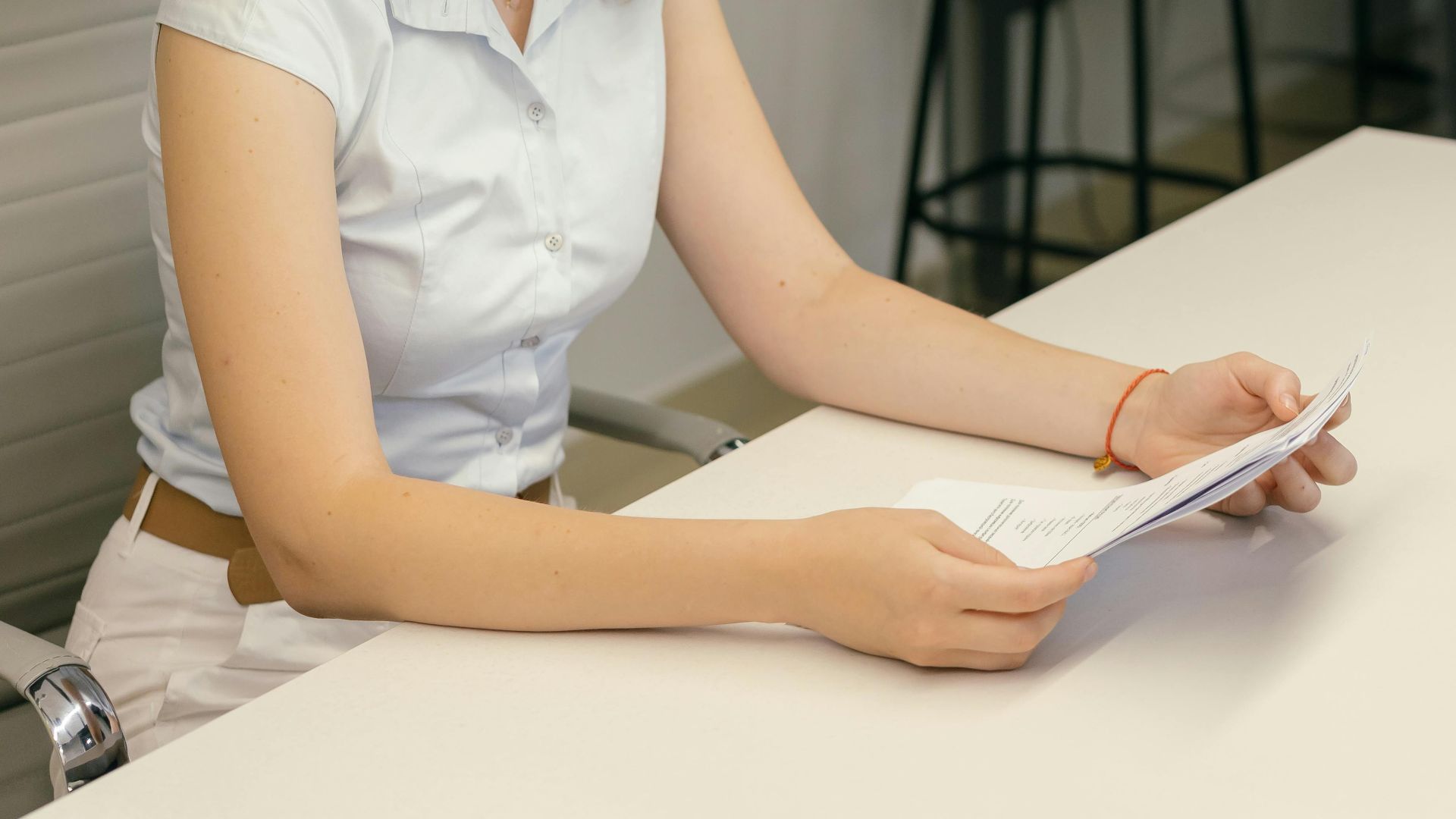 A woman in a modern office reading documents at a desk beside an indoor plant.