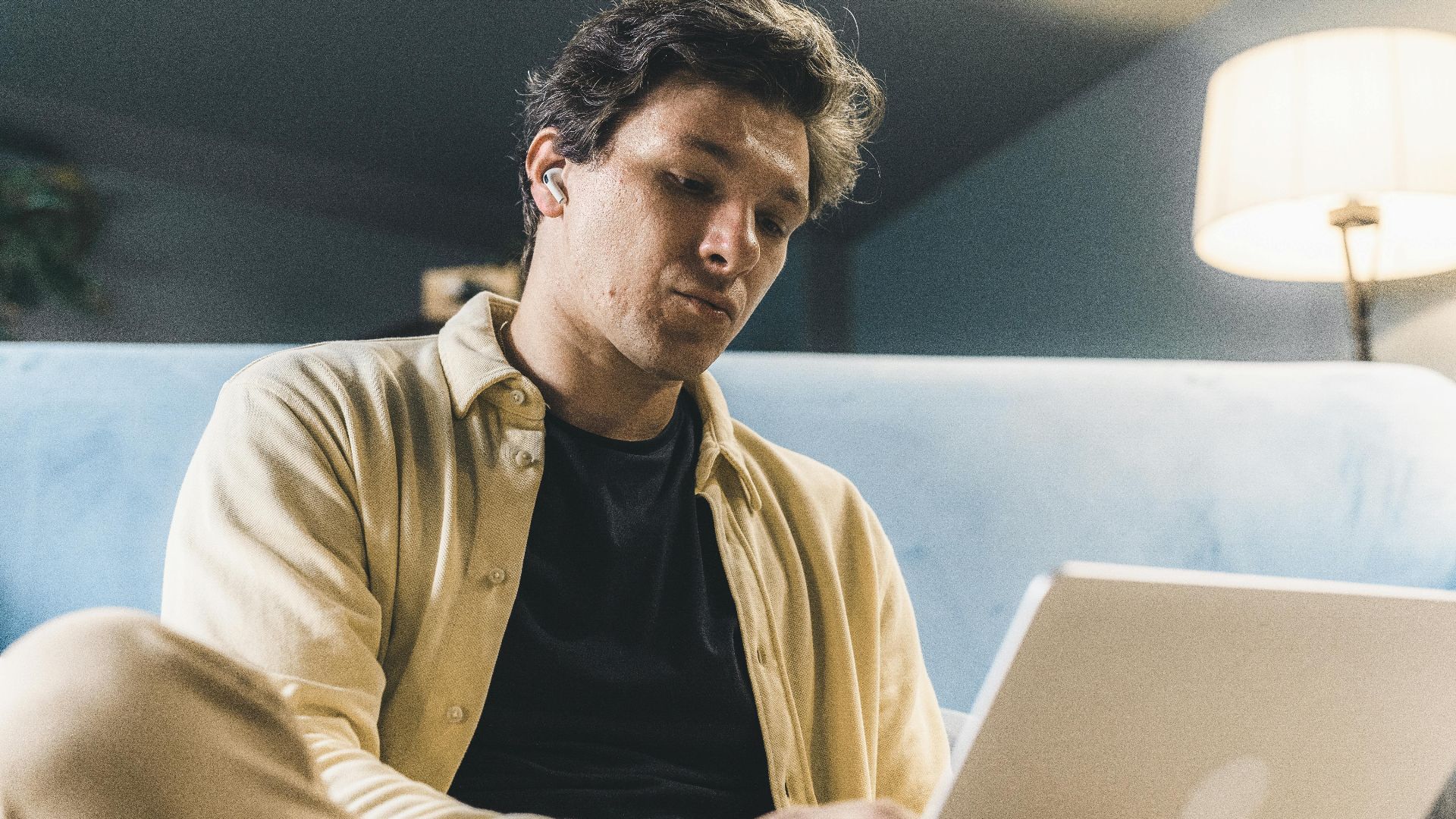 A young man sits indoors, working on his laptop with a thoughtful expression, under warm lighting.