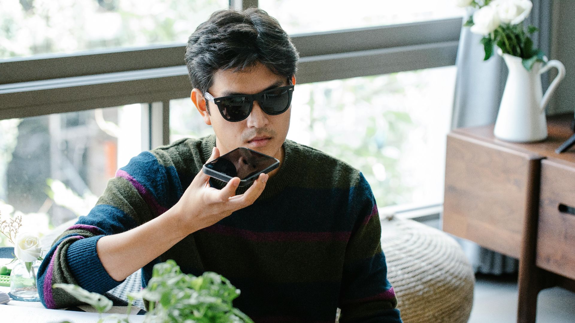 A man wearing sunglasses uses a smartphone's voice assistant in a cozy indoor setting.
