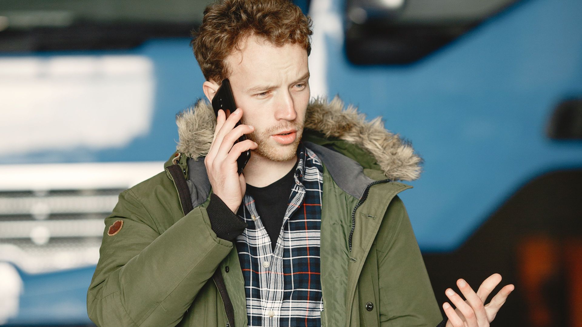 Man wearing green jacket and plaid shirt talks on the phone near a truck outdoors.