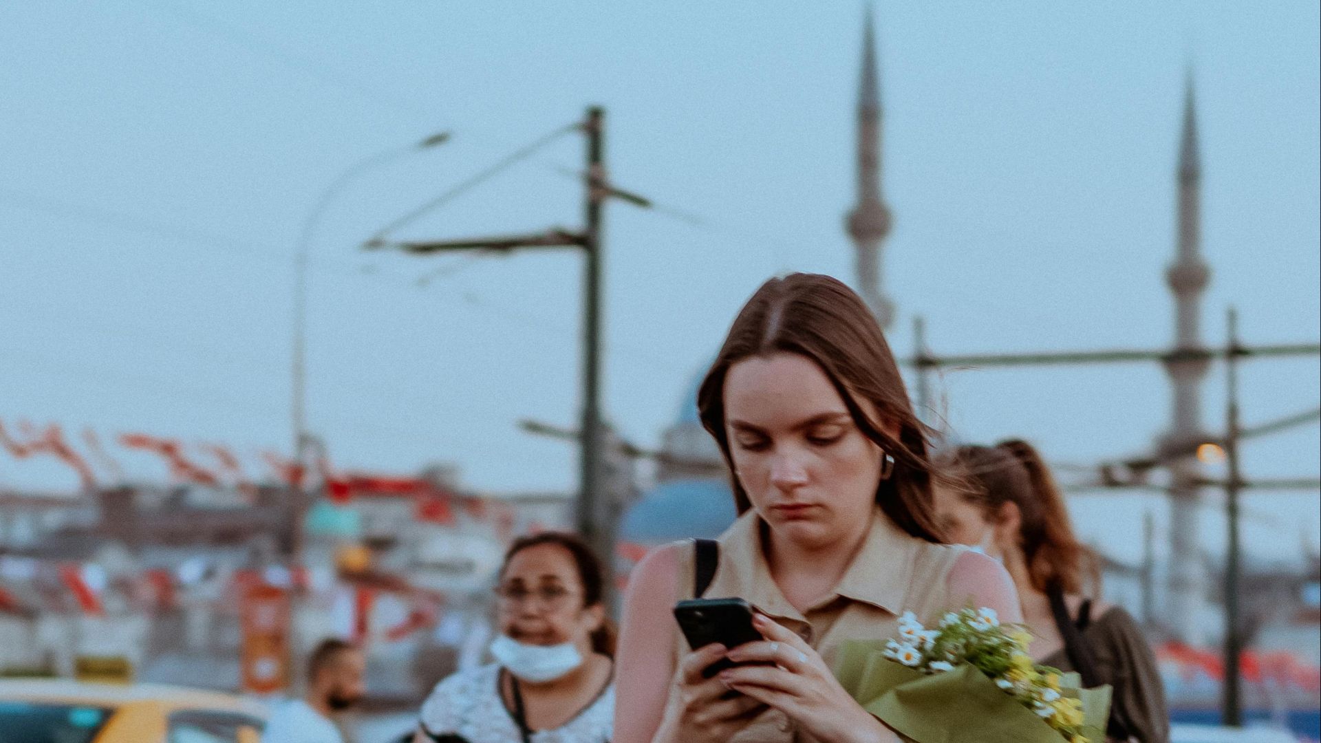 A young woman carrying flowers and using a smartphone while walking on a busy city street.