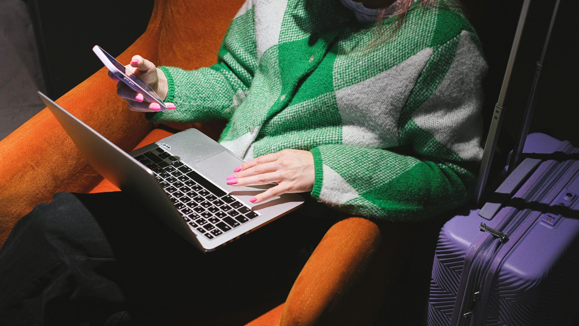 Woman in a knitted sweater working remotely on laptop with a smartphone, sitting in an armchair.