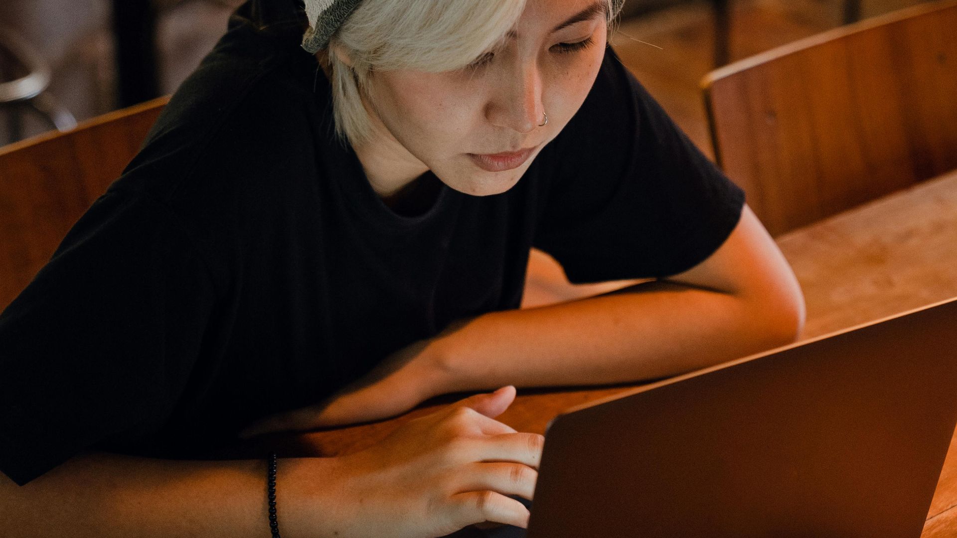 From above of young Asian lady with blond bangs in slouchy beanie and black T shirt sitting at table and browsing Internet sites on laptop in modern bar