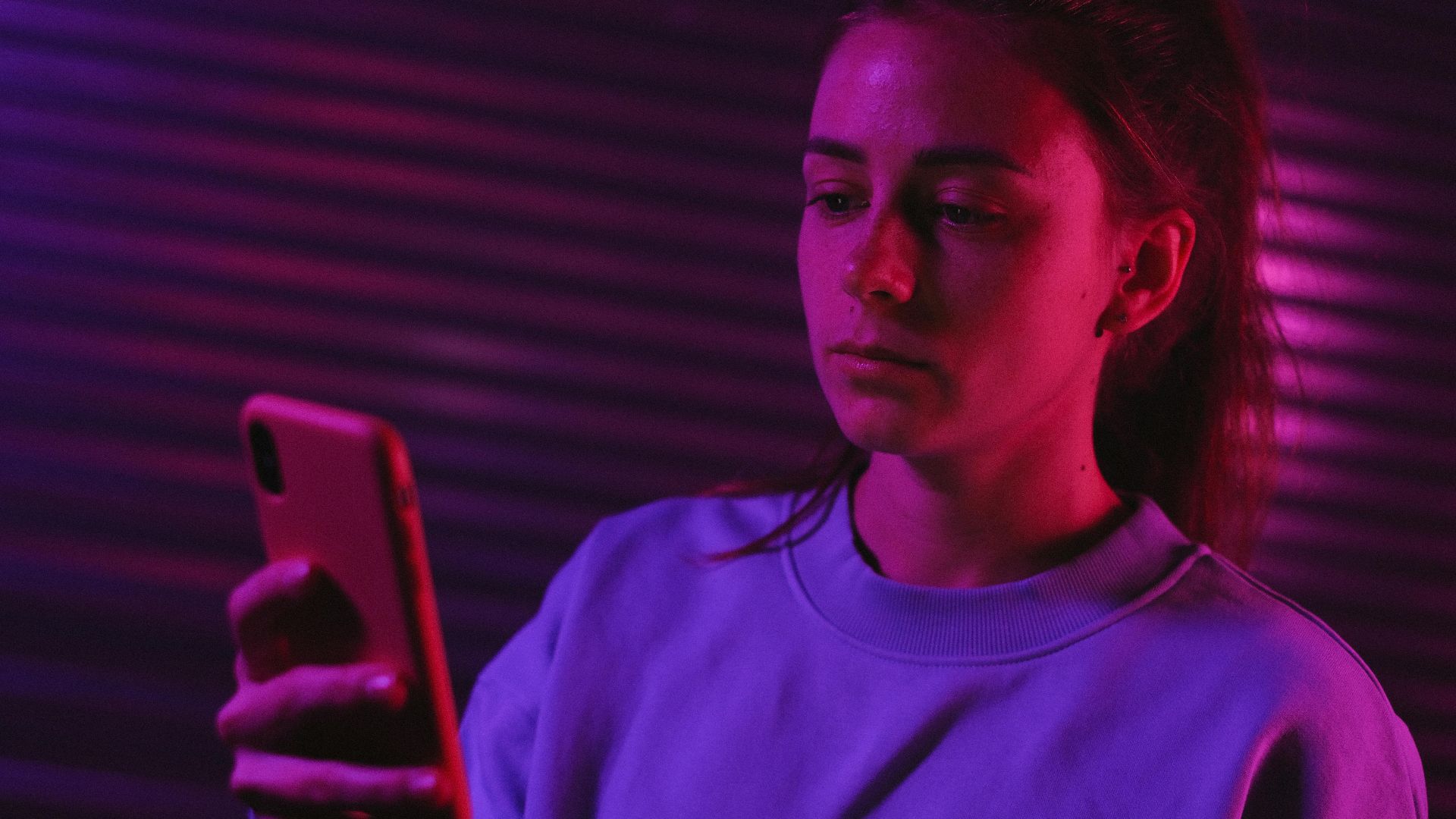 Thoughtful young woman in casual clothes using smartphone in dark room with neon purple light