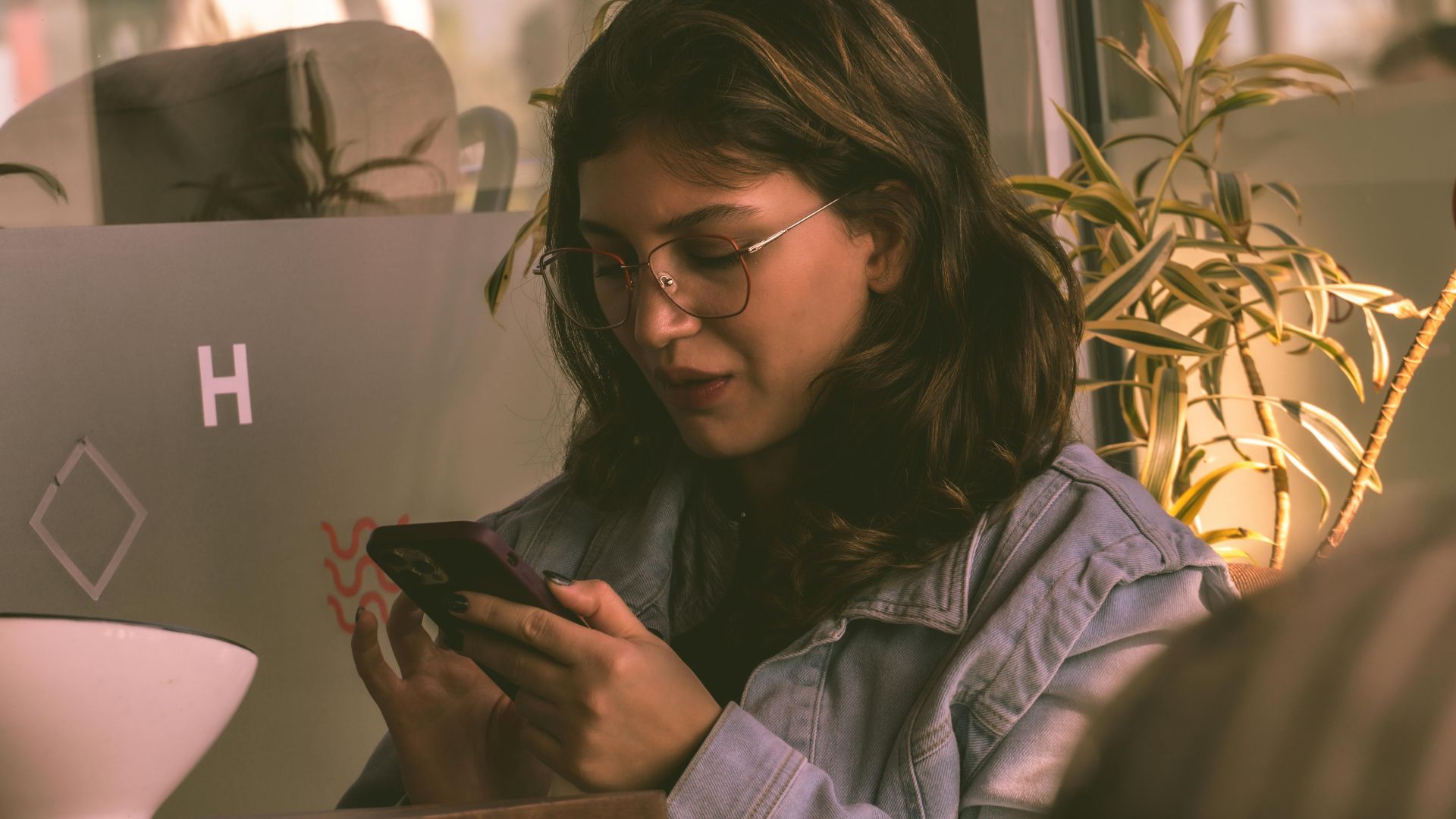 A young woman wearing glasses and denim jacket using a smartphone in a cozy Istanbul cafe.