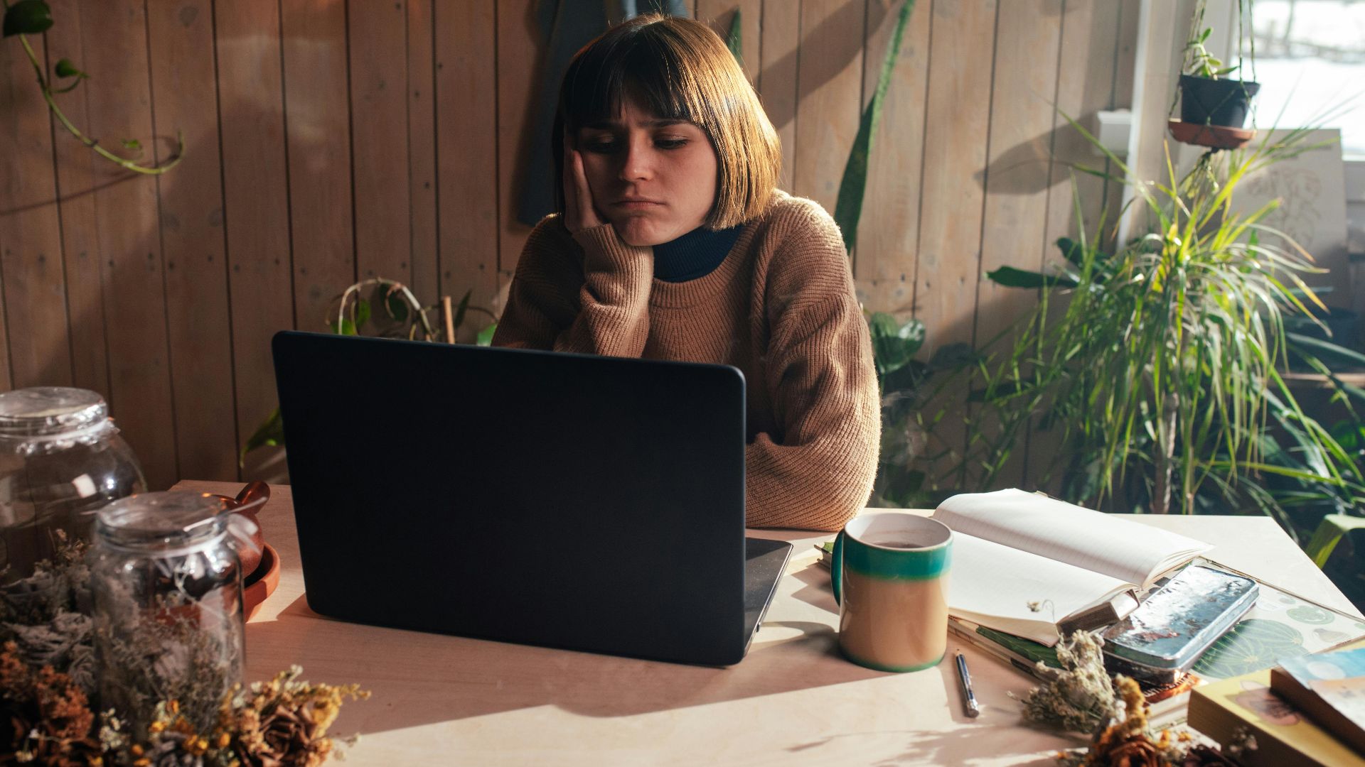 A woman in a cozy room surrounded by plants, appearing bored while using her laptop.