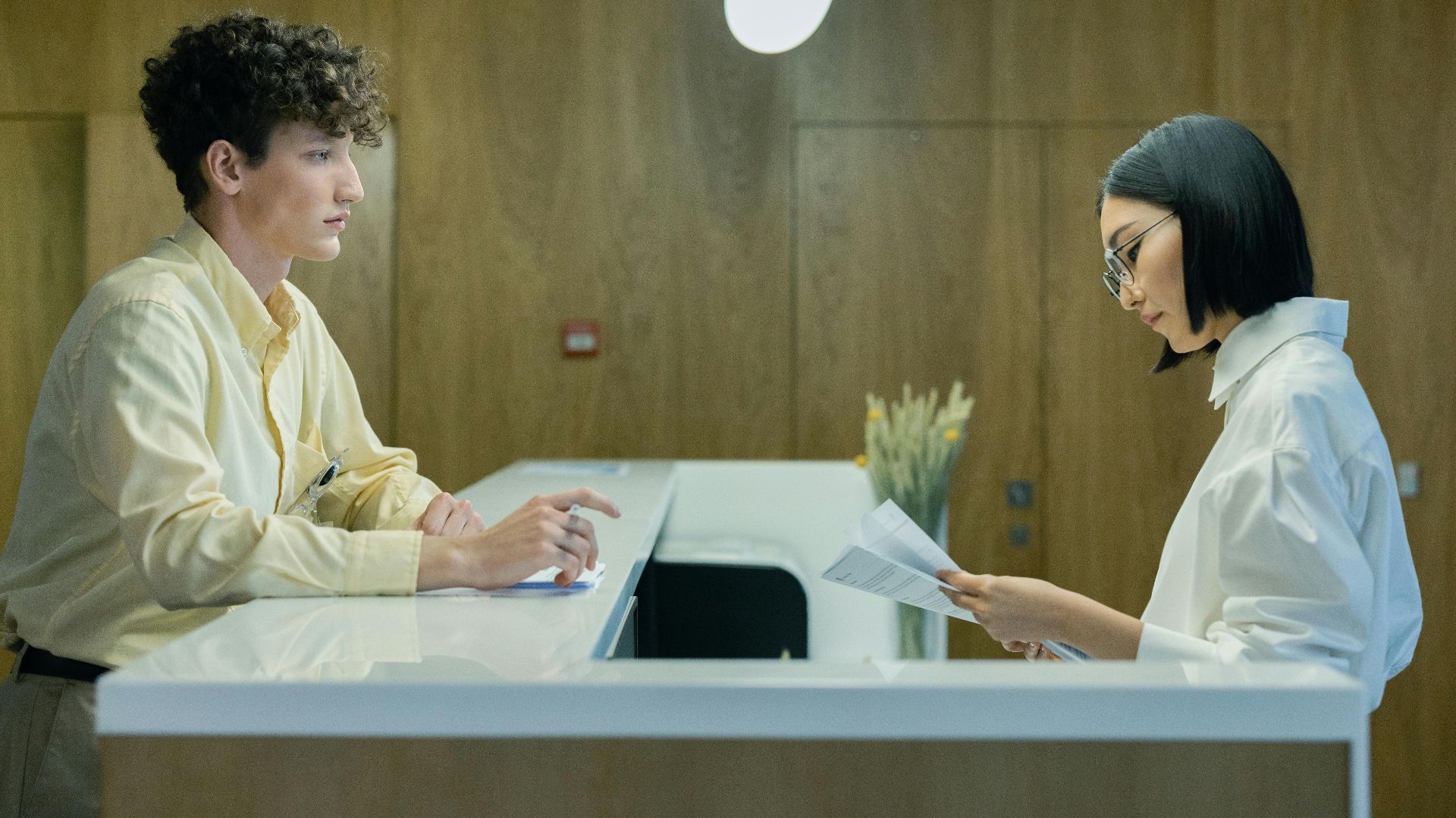 A young man and woman interacting at a modern office reception desk indoors.