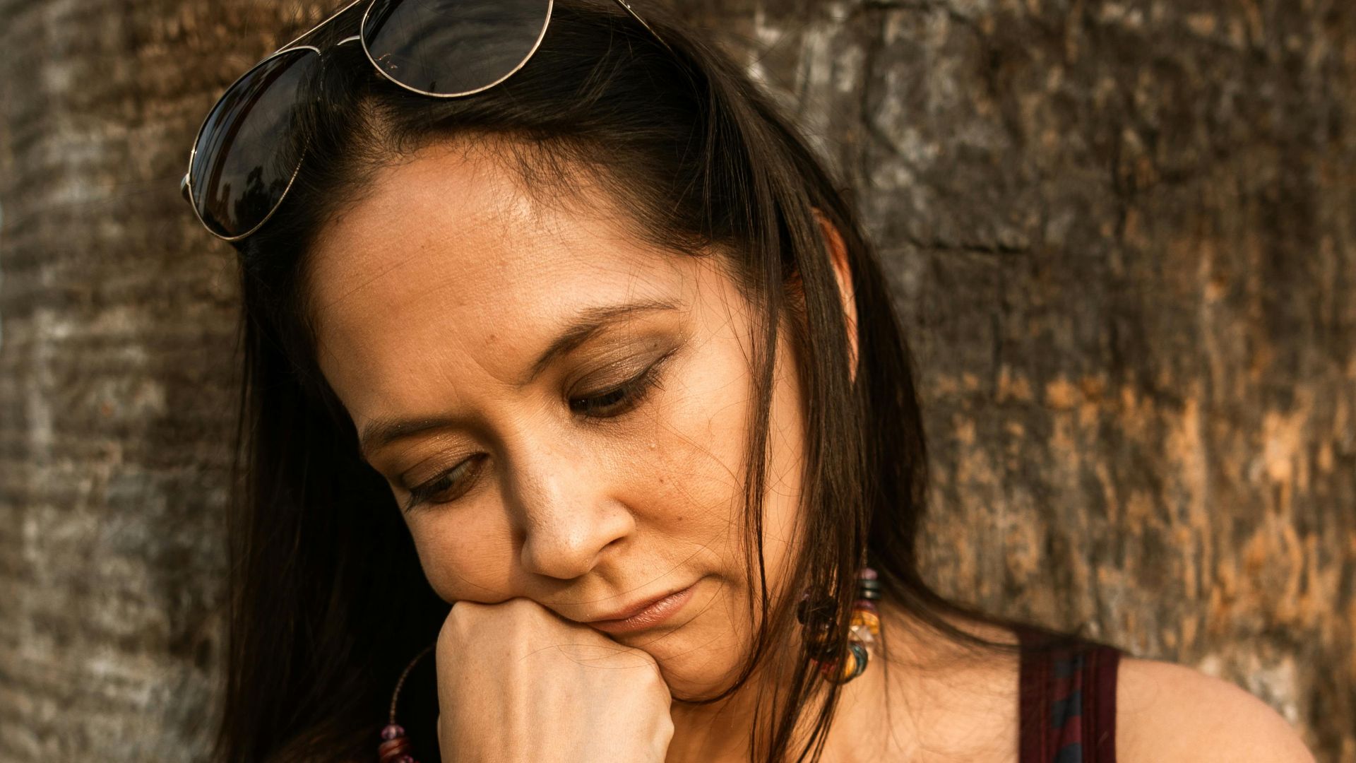 Woman in striped tank top leaning against tree, looking contemplative and pensive.