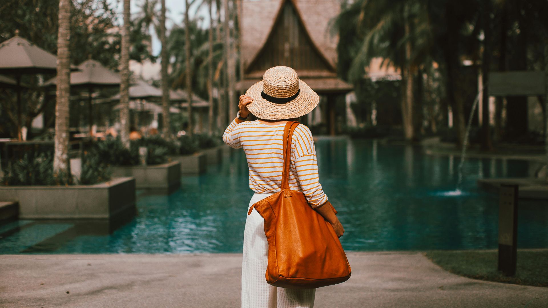Back view faceless female in summer wear and hat standing on poolside in tropical resort