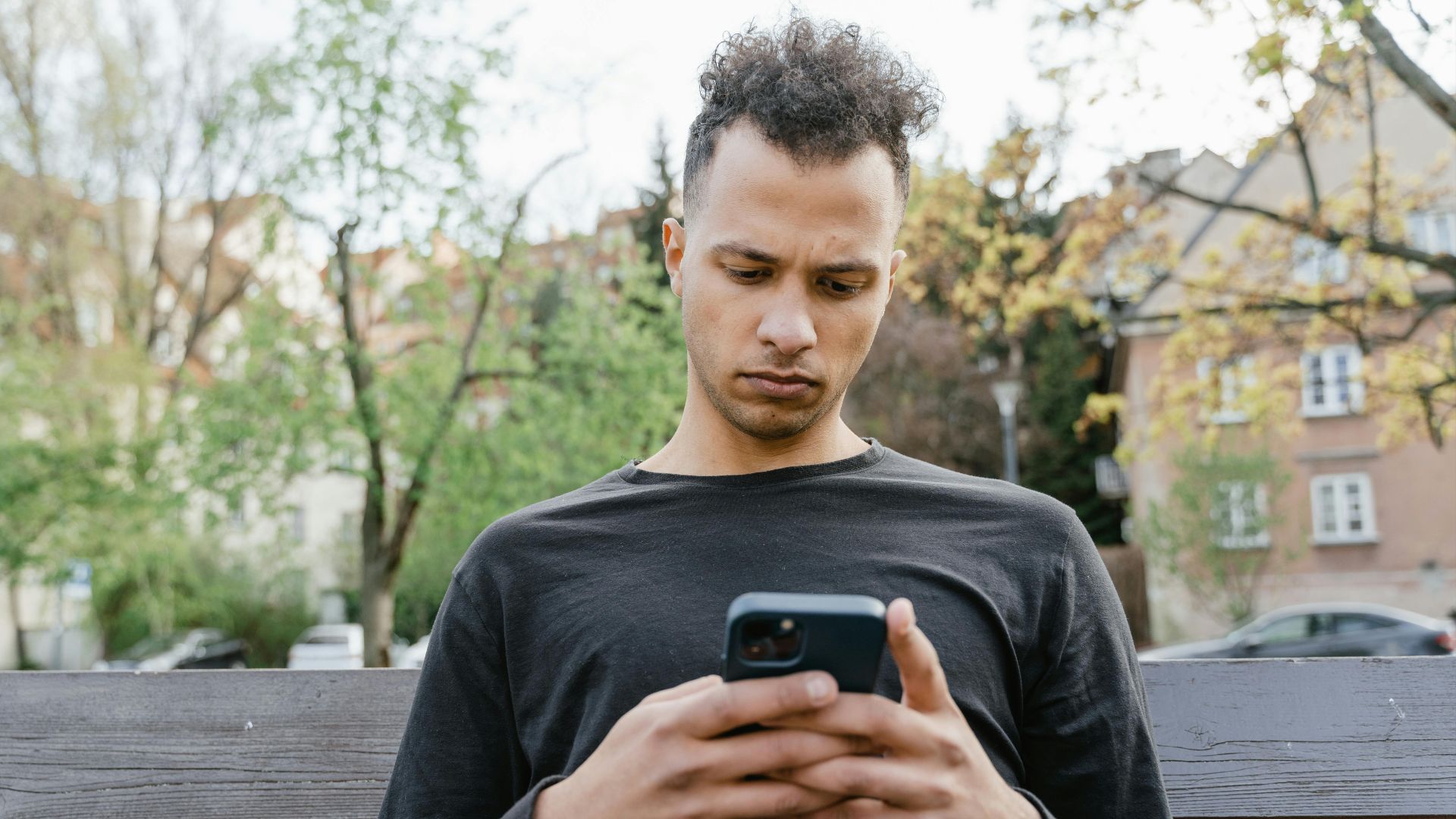Casual young man sitting on a bench, texting on his smartphone in a park setting.