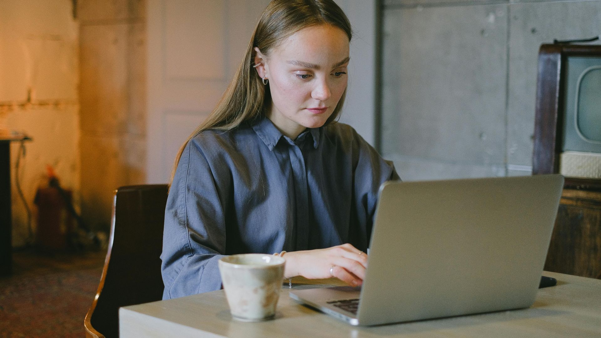 Concentrated young lady in casual outfit sitting on chair at table and working remotely on laptop near cup of drink in loft