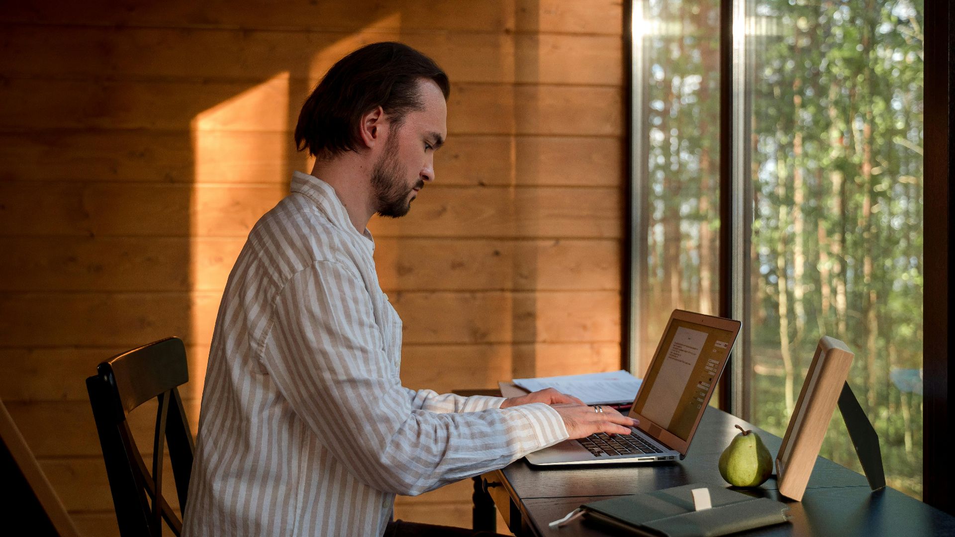 Side view of a man with facial hair typing on a laptop indoors by a window, illuminated by soft, natural light. Modern home office setup with books and a notebook on the desk.