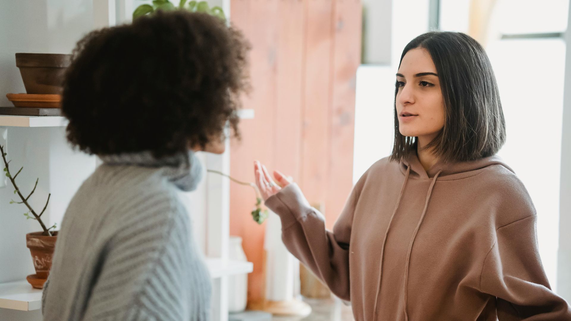 Serious woman in casual wear having argument with anonymous female with curly hair while standing in light room at home