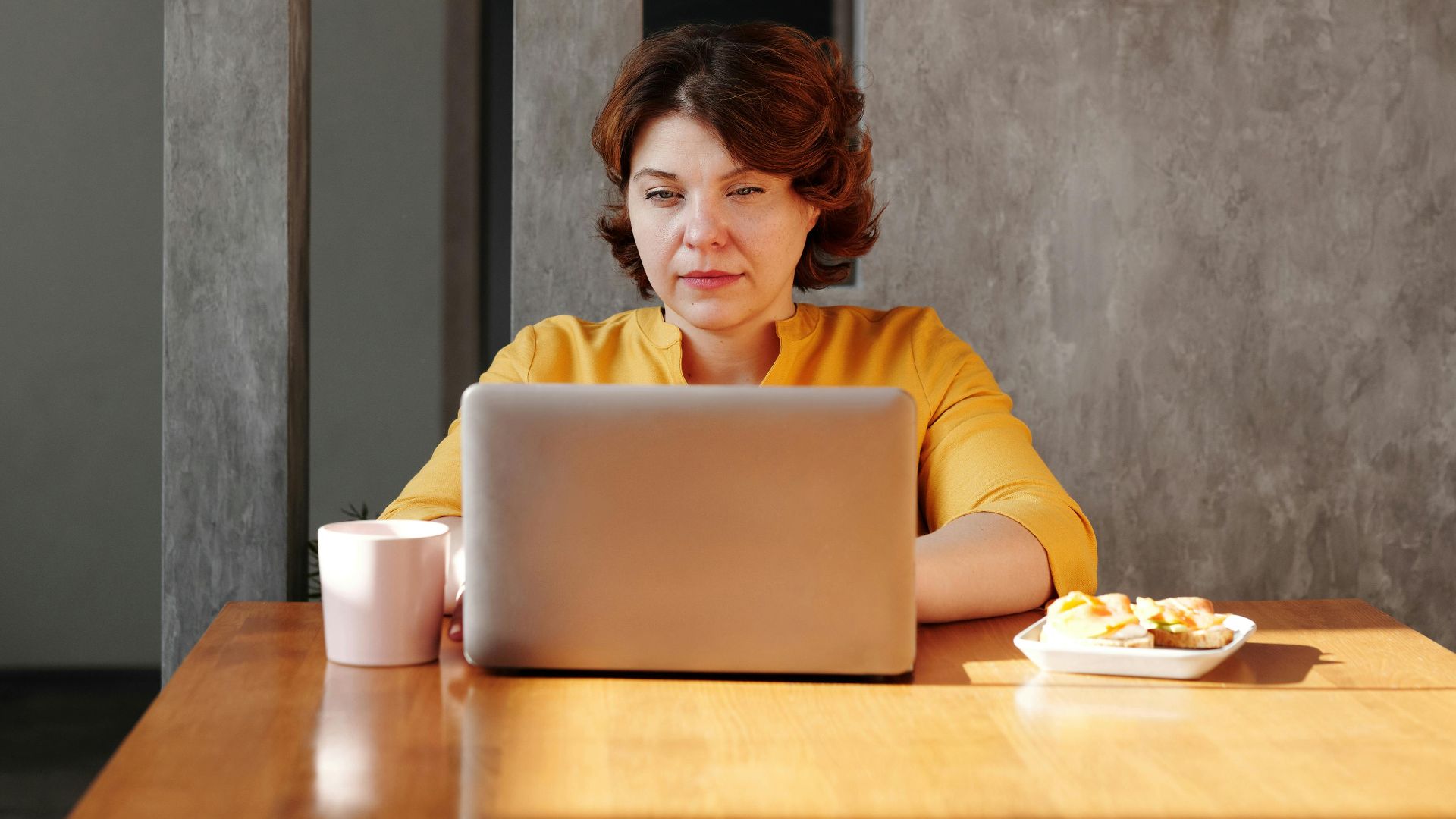 Woman using a laptop at a wooden table with coffee and snacks, embodying modern remote work lifestyle.