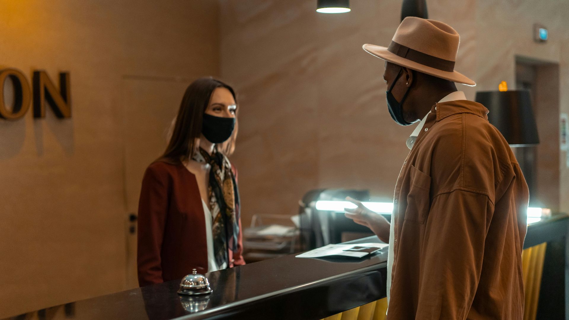 A man at a hotel reception talking to a staff member. Both wear face masks.