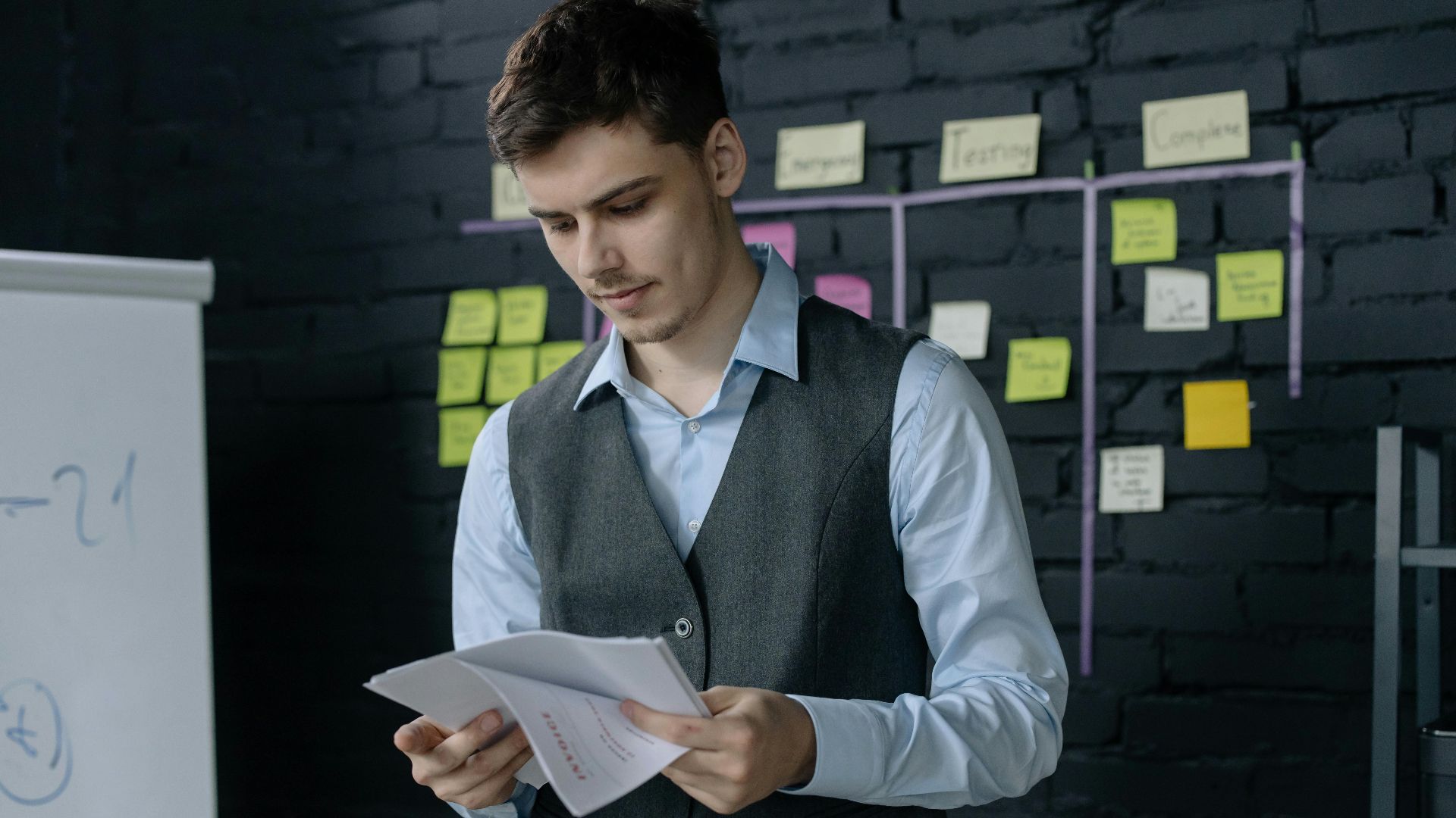 A young man reviewing documents in an office with a black brick wall and sticky notes background.