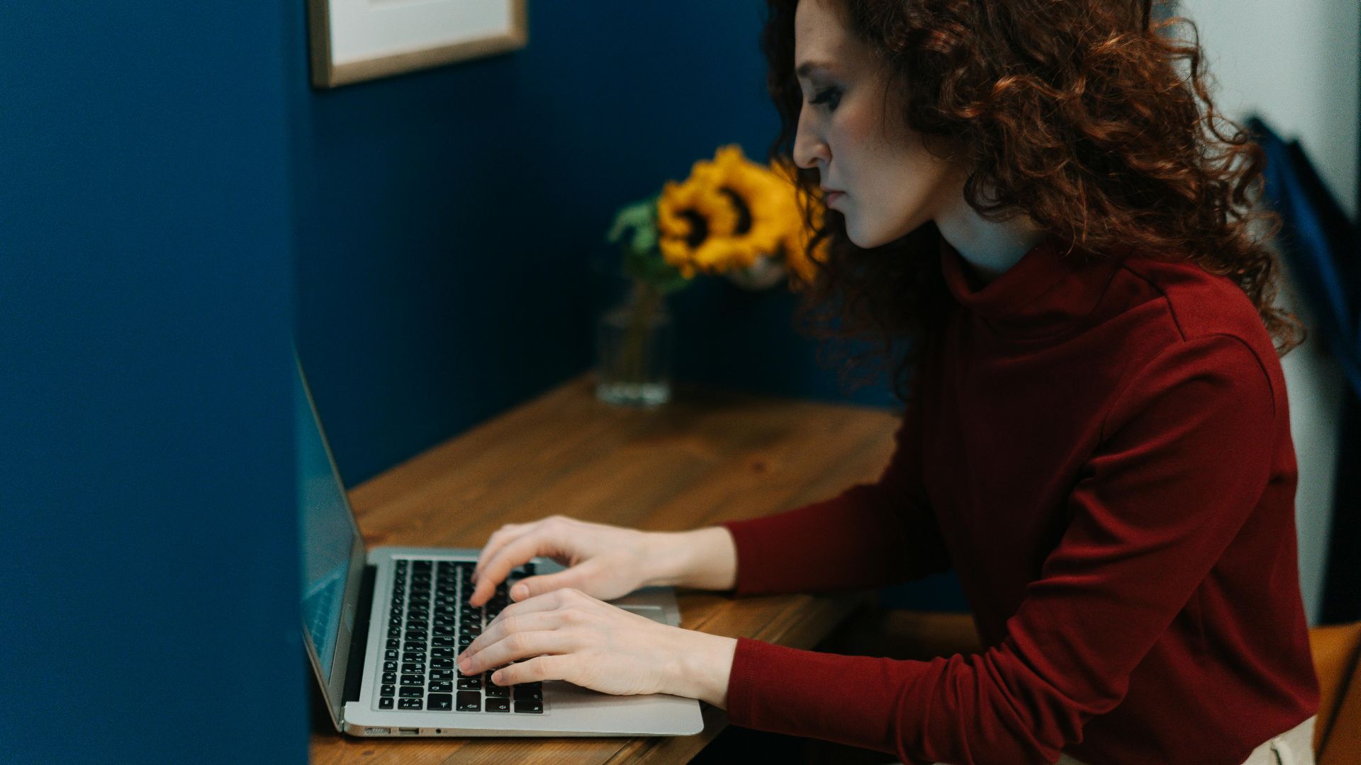 A woman working on a laptop at a desk with sunflowers, surrounded by blue decor, focusing intently.