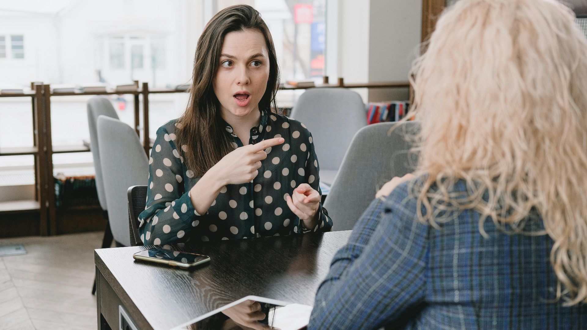 Astonished female pointing away while sitting at table with smartphone and tablet and talking to woman