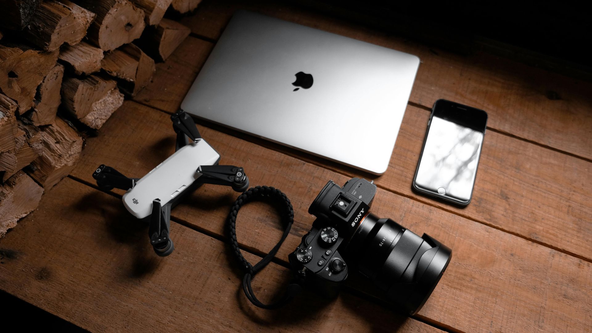 A flat lay of modern technology devices including a laptop, camera, drone, and smartphone on a wooden table.