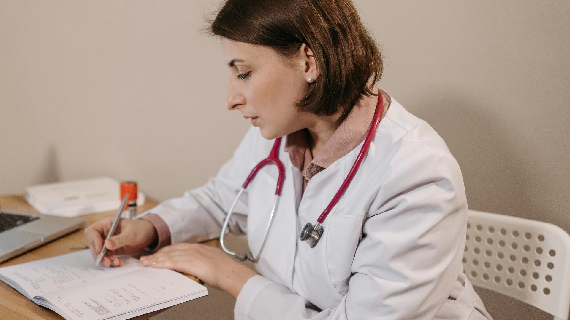 A female doctor in a lab coat writing a prescription in a clinic office setting with a laptop on the table.