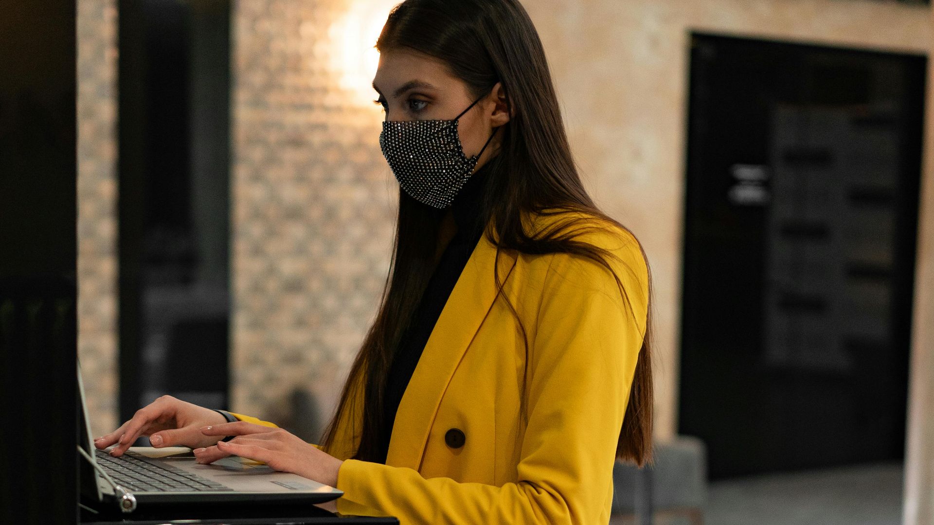 Woman wearing mask and yellow coat using laptop in hotel lobby next to suitcase.