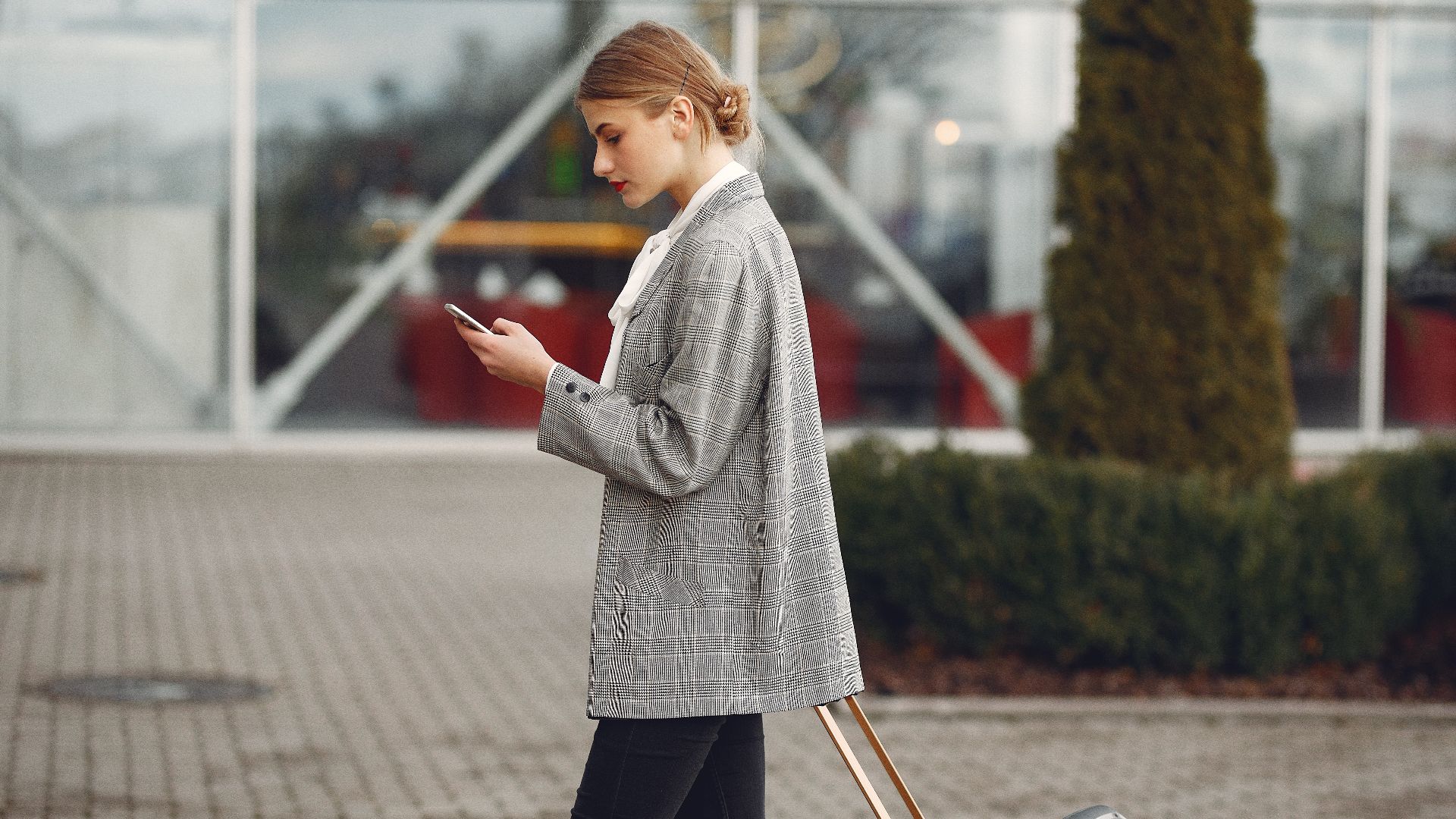 Stylish woman walking with suitcase while using smartphone outside a modern building.