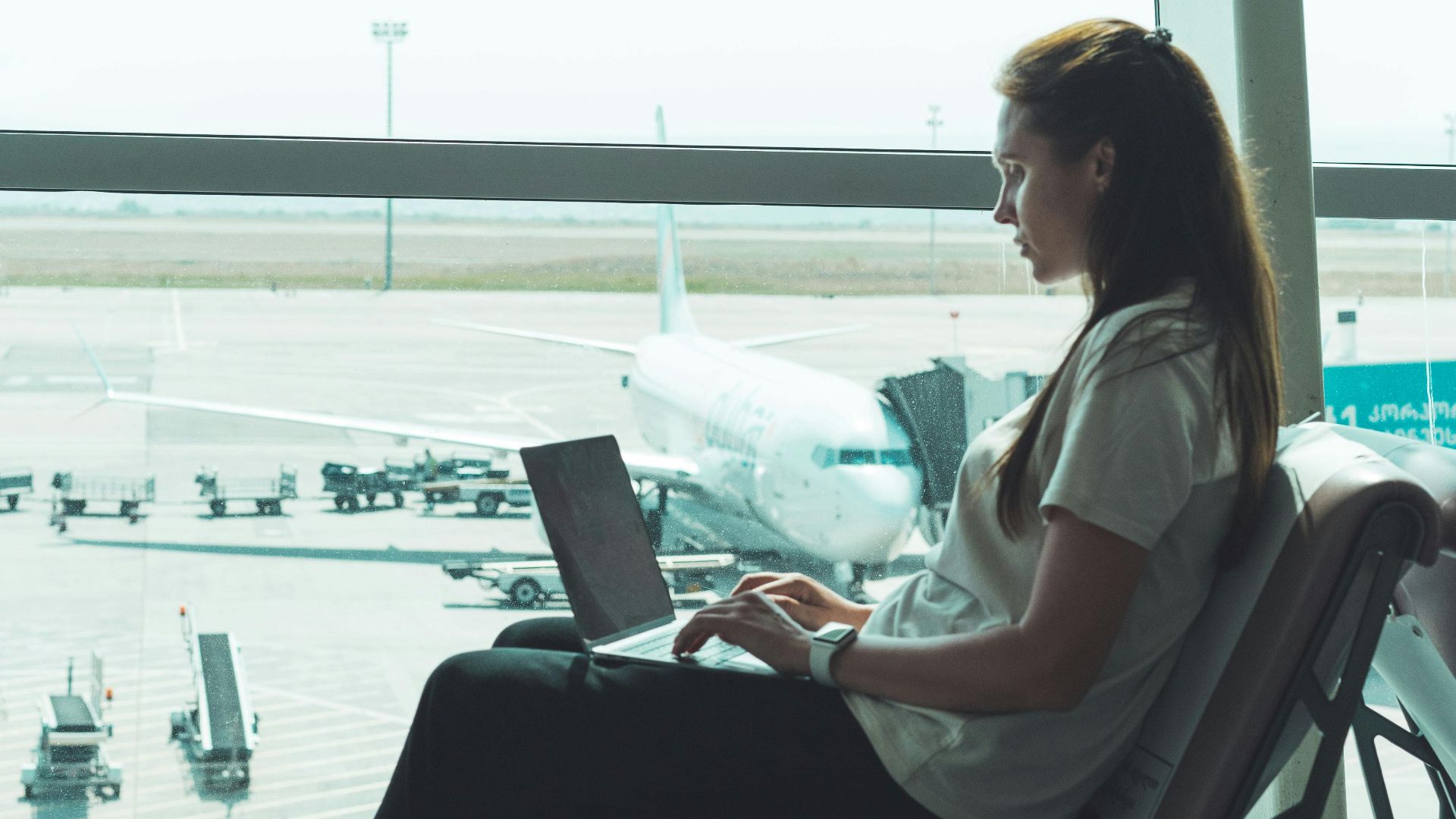 Side view of woman using laptop at airport, airplane visible outside through large glass windows.