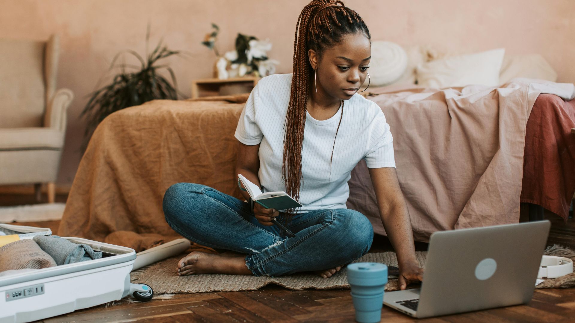 Woman sits on floor packing suitcase, using laptop, and holding notebook in a cozy bedroom environment.