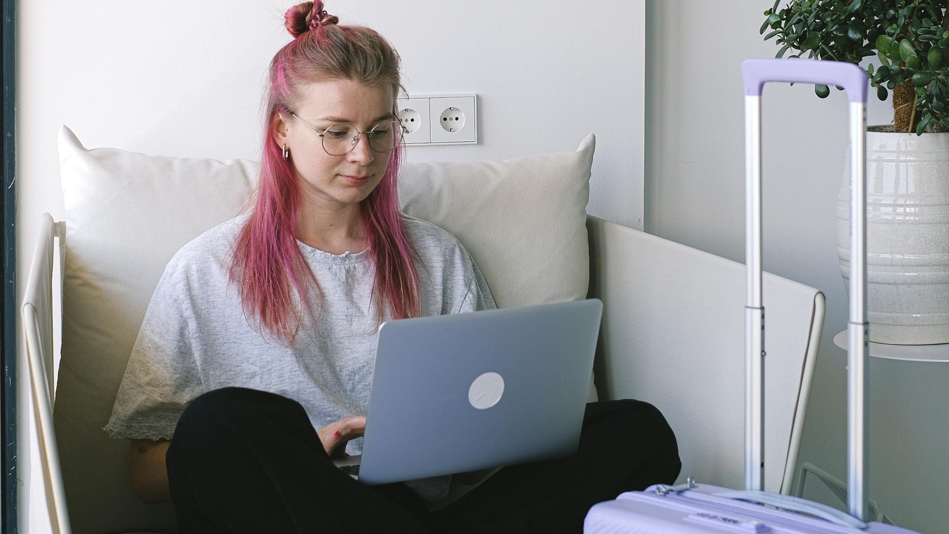 Woman with pink hair working on a laptop while sitting on a sofa in a hotel room with luggage.