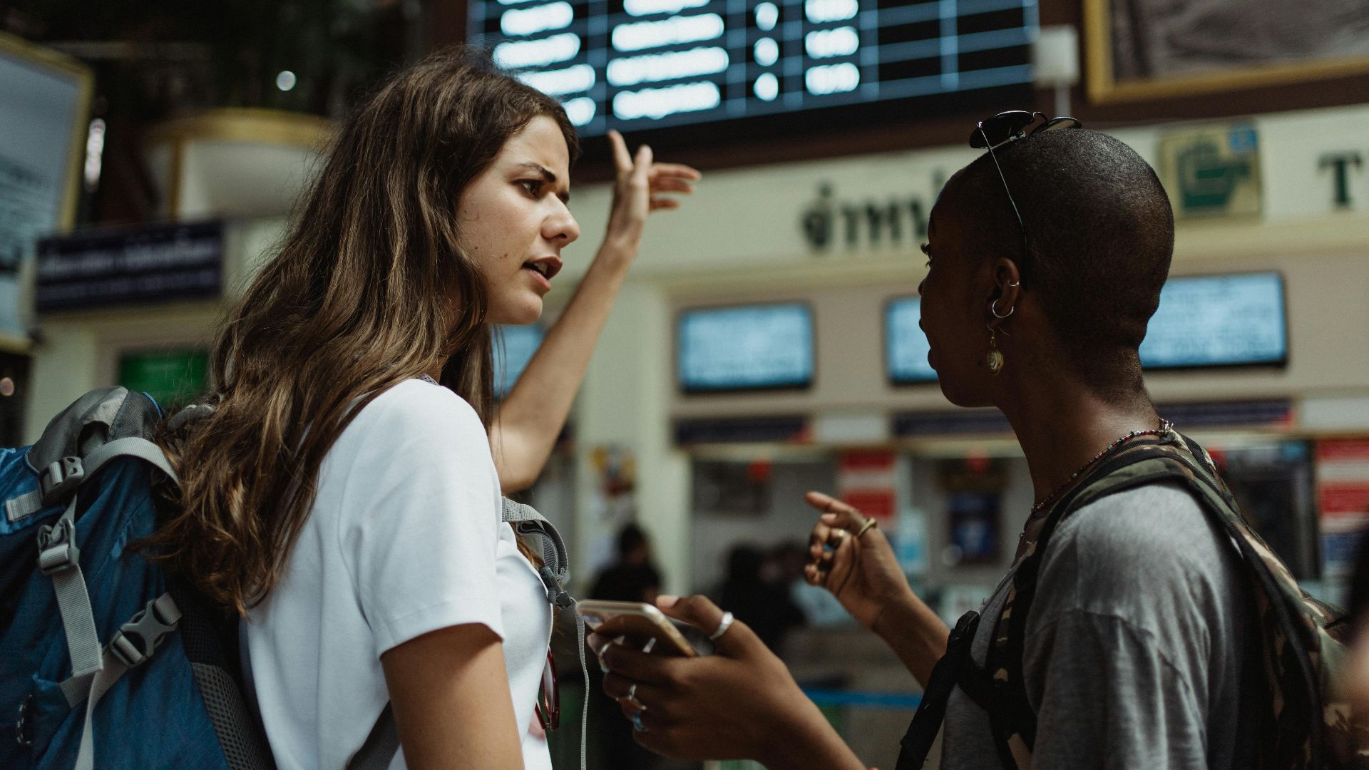 Two travelers with backpacks discuss in front of an airport timetable. Adventure awaits.