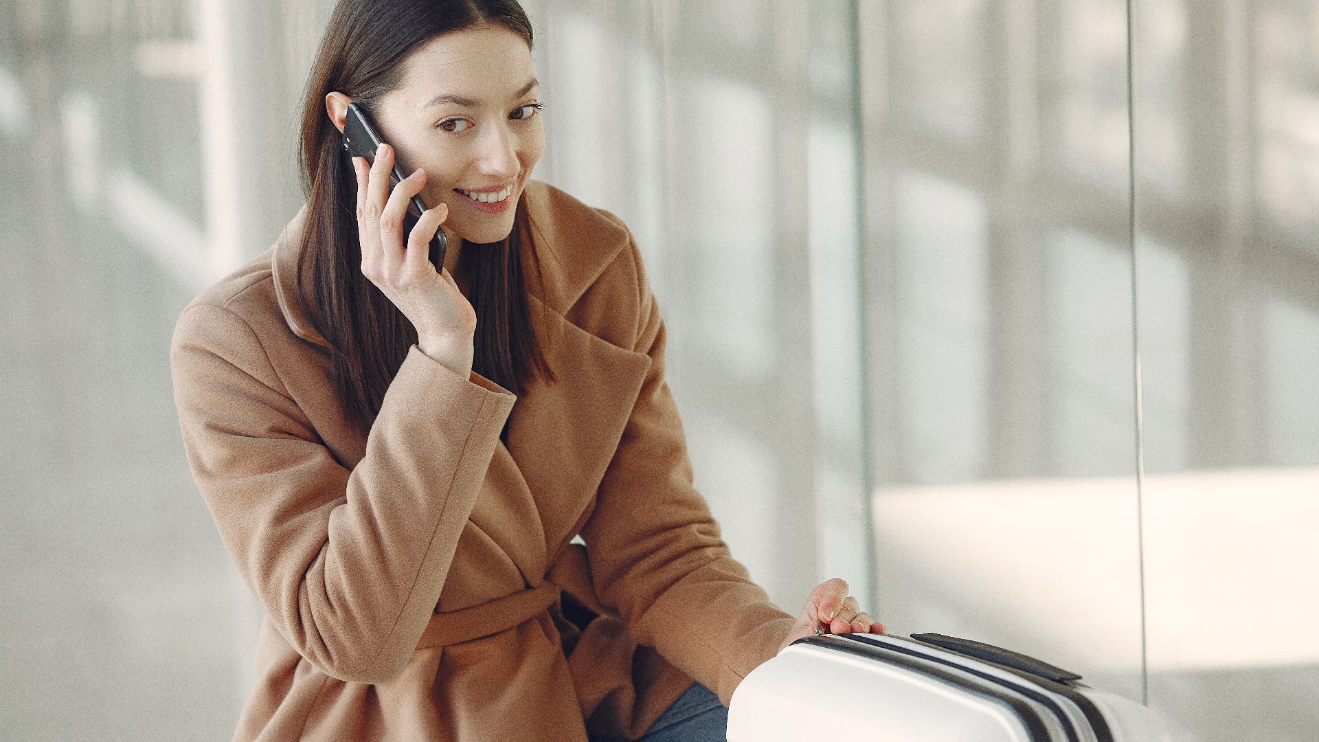 Young dreamy brunette in beige coat with suitcase having conversation via mobile phone while waiting for flight in airport terminal