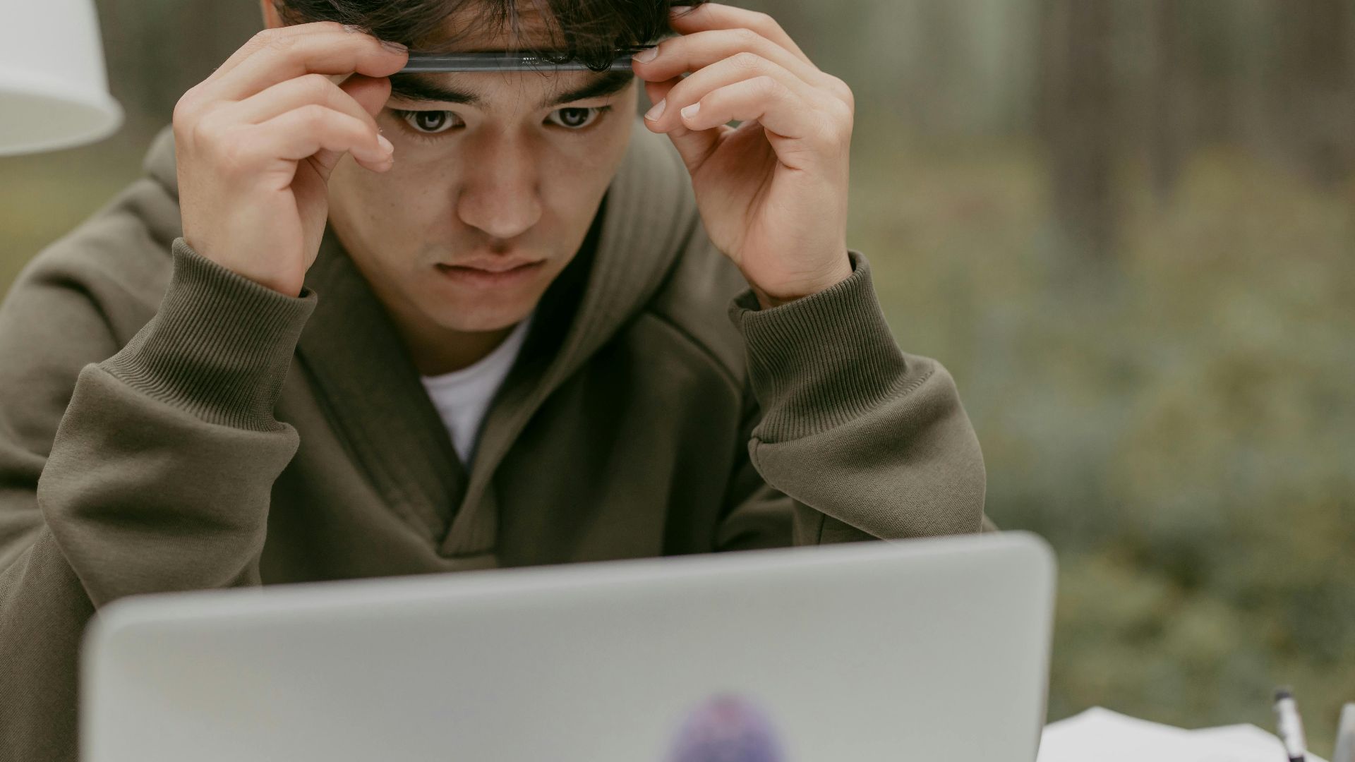 A young man in a green jacket concentrating on a laptop screen in an outdoor setting.