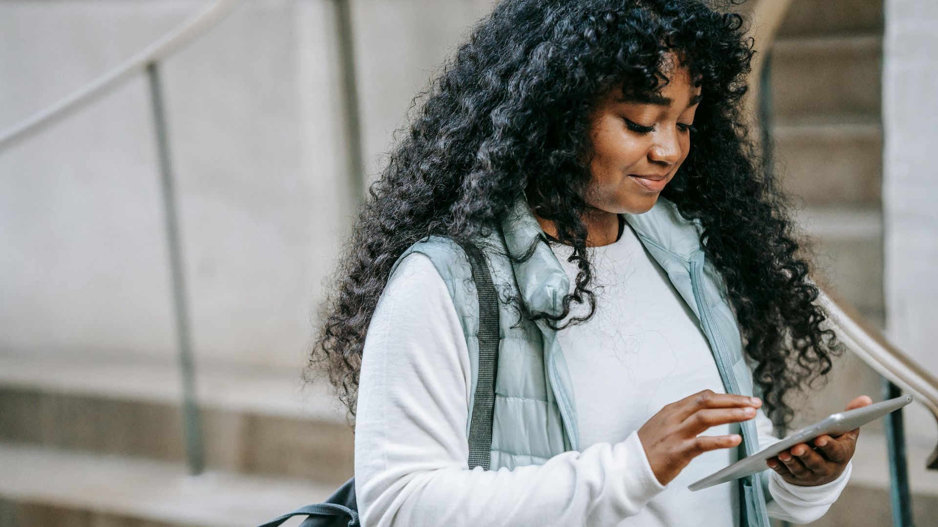 Content African American female with long curly hair wearing warm jacket browsing modern tablet while standing on city street