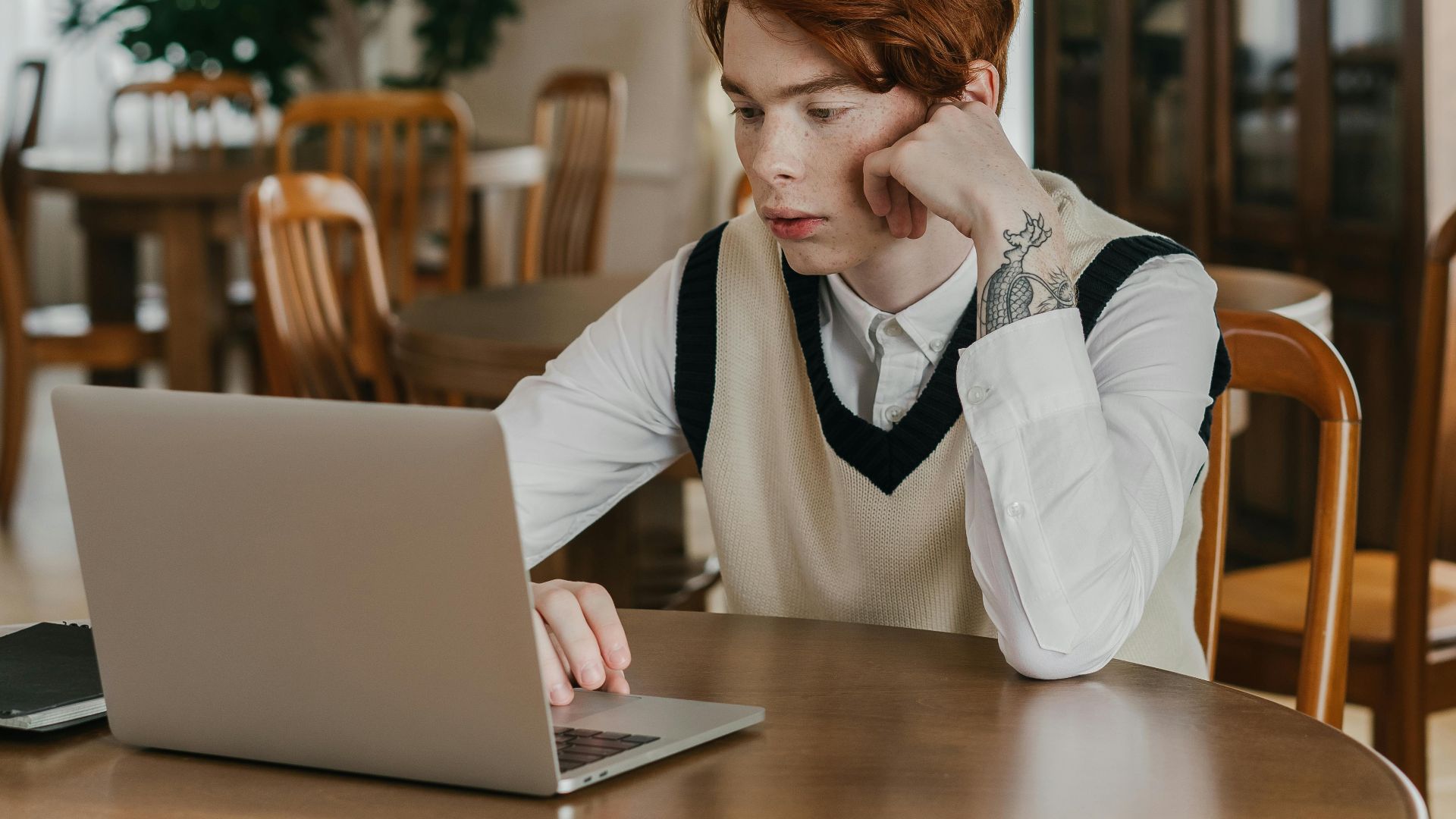 A young adult focused on his laptop while seated at a round table in a cozy cafe environment.