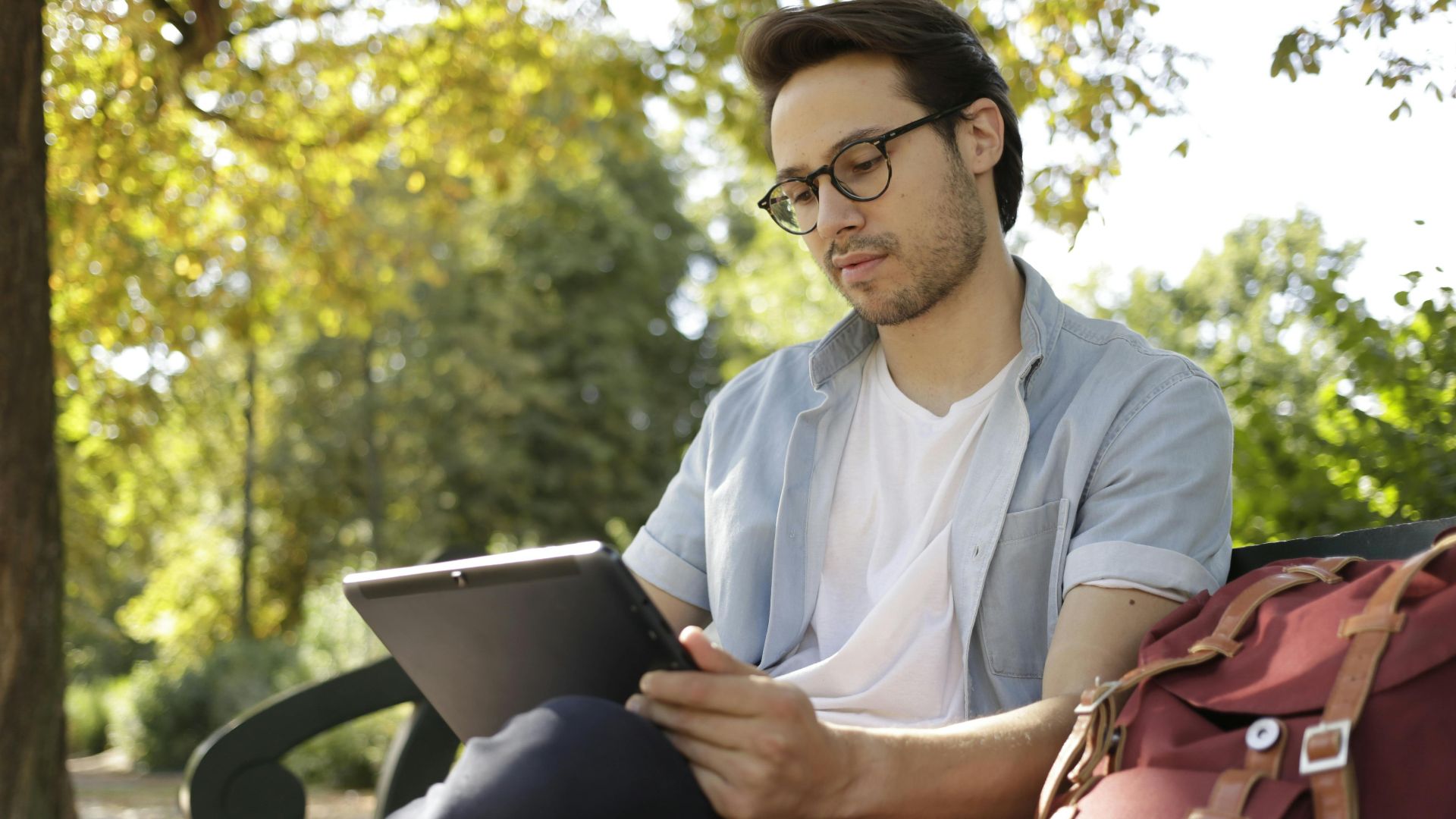Relaxed young man reading a tablet outdoors in a sunny park setting.