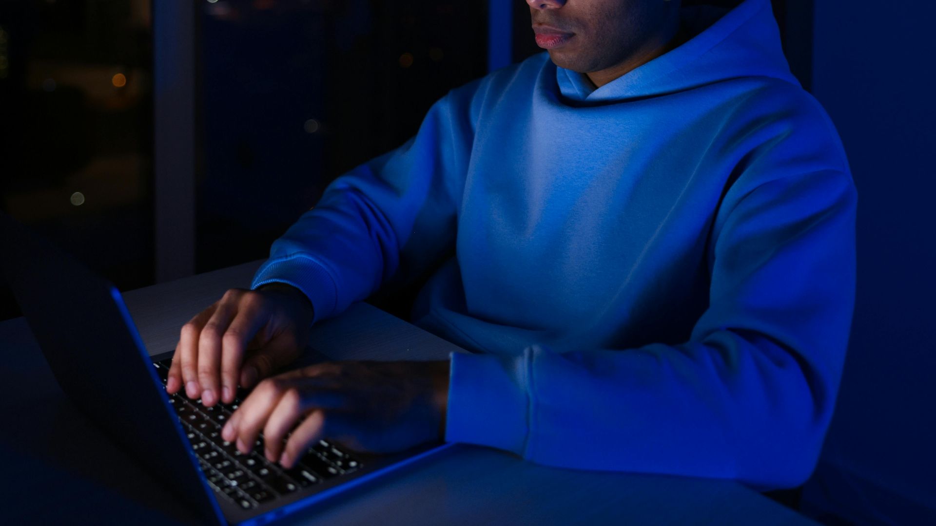 Young man in hoodie typing on laptop indoors at night, illuminated by screen light.