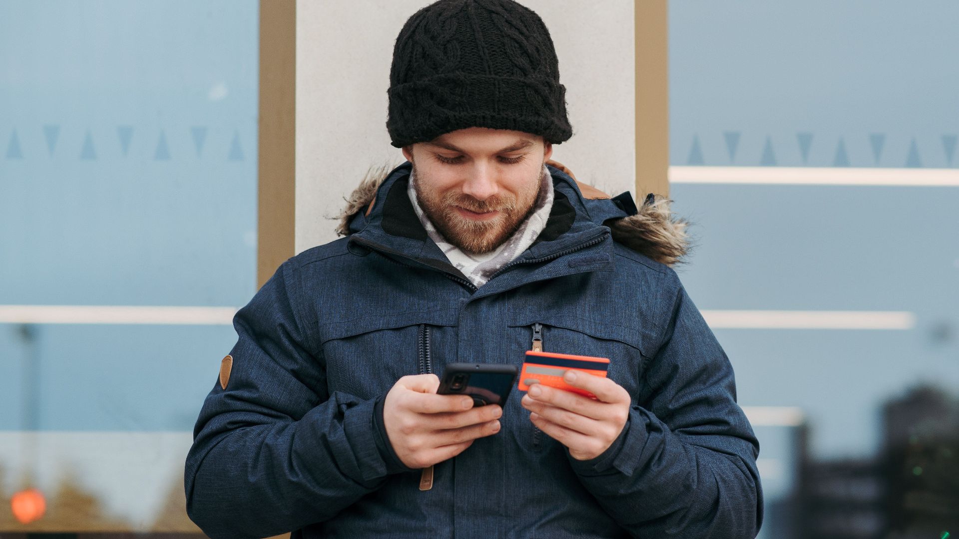 Positive man in warm clothes standing near building and entering credentials of credit card on mobile phone in daylight