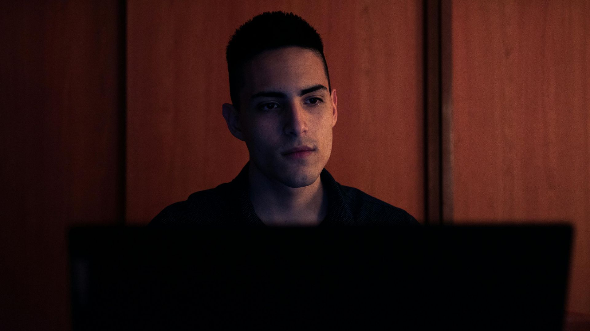A young man working on his laptop, focused and serious, in an indoor setting with warm lighting.