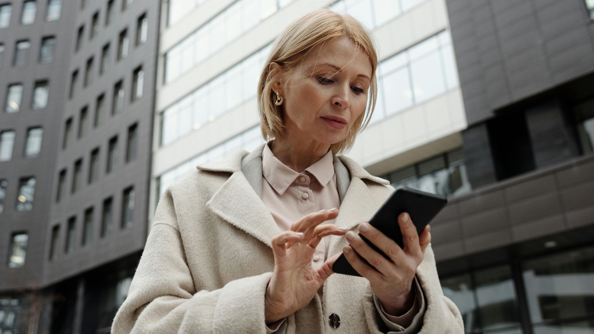 Woman in coat using smartphone in front of modern building. Professional and focused expression.