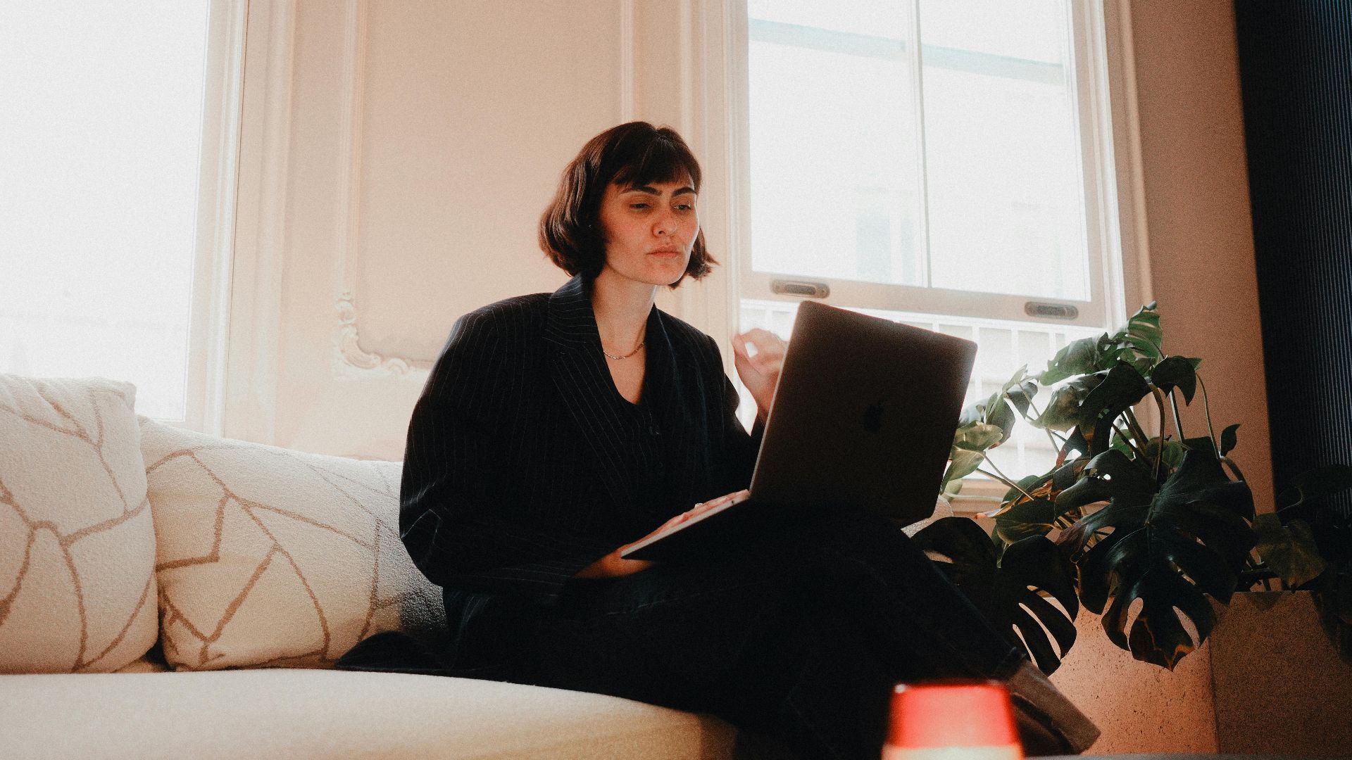Woman in suit using laptop on sofa in bright room, modern lifestyle.