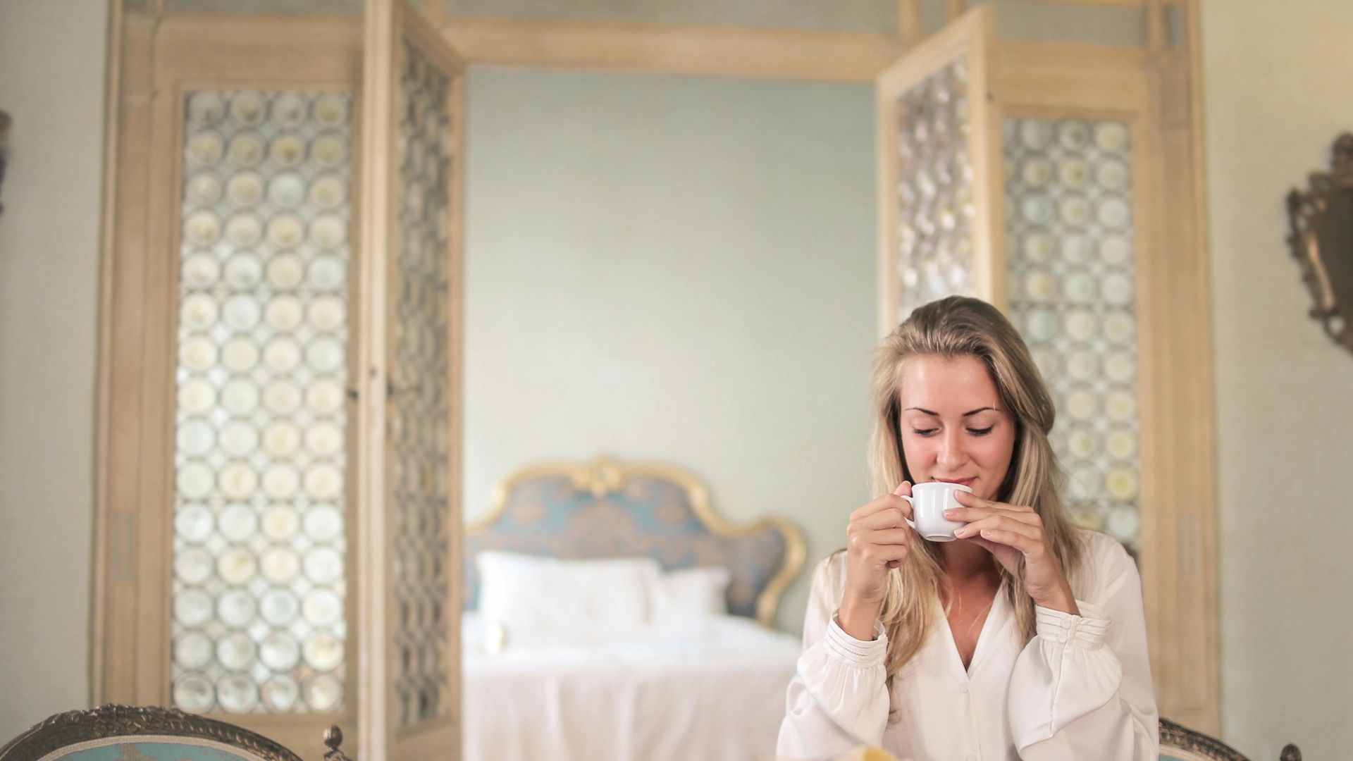 Glad female in elegant wear sitting at table with metal tray of healthy breakfast in vintage hotel room and drinking coffee in morning