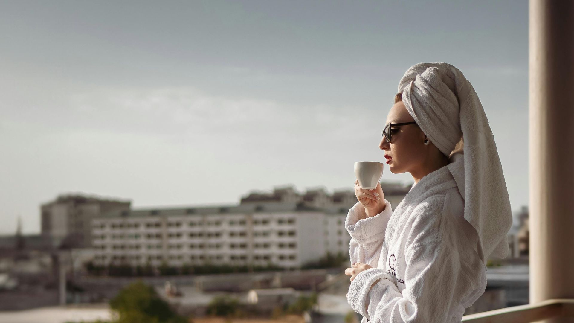 A woman in a bathrobe enjoys a drink on a sunny balcony overlooking the ocean.