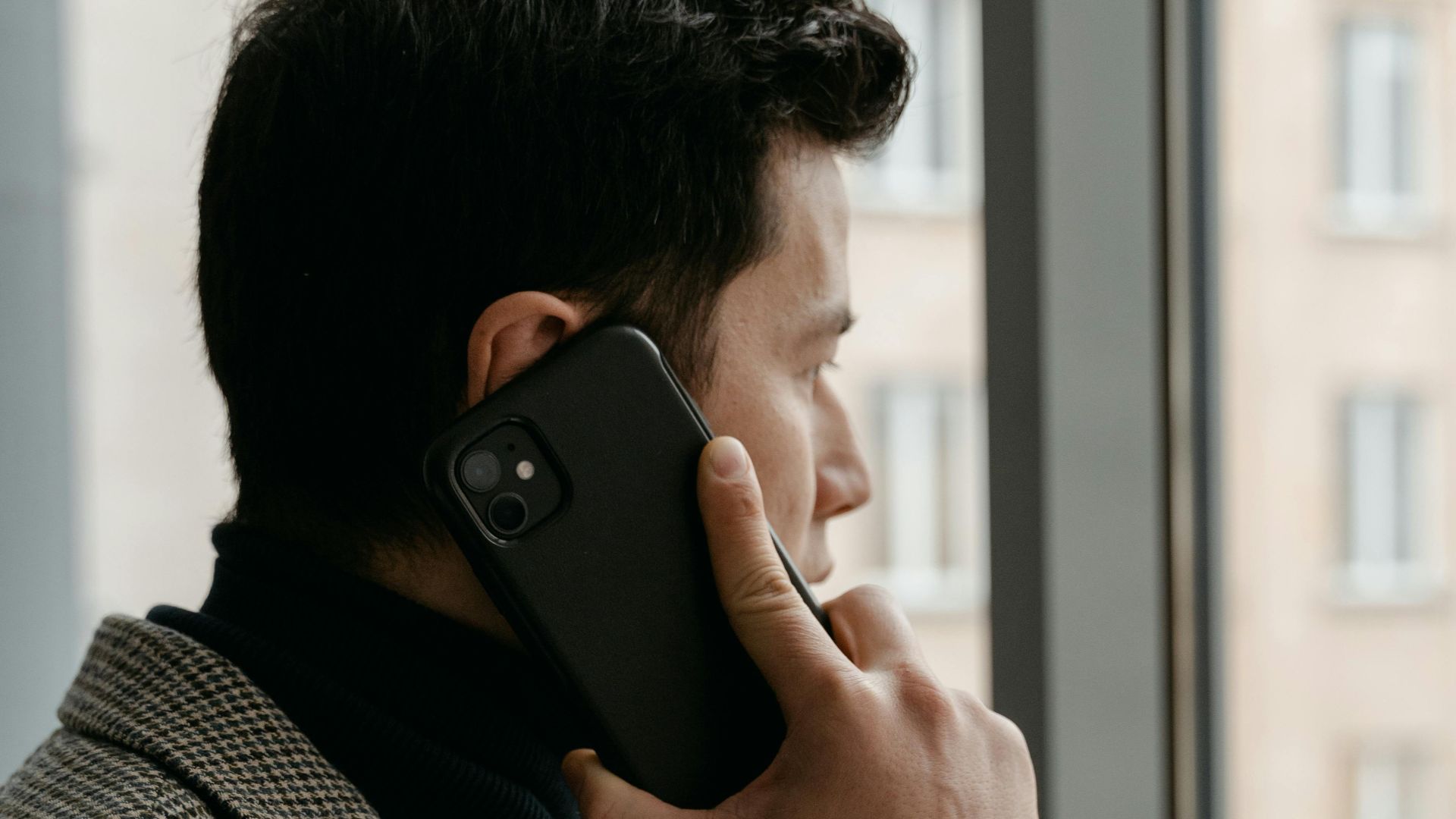 Man in a checkered blazer using a smartphone while gazing out a window.
