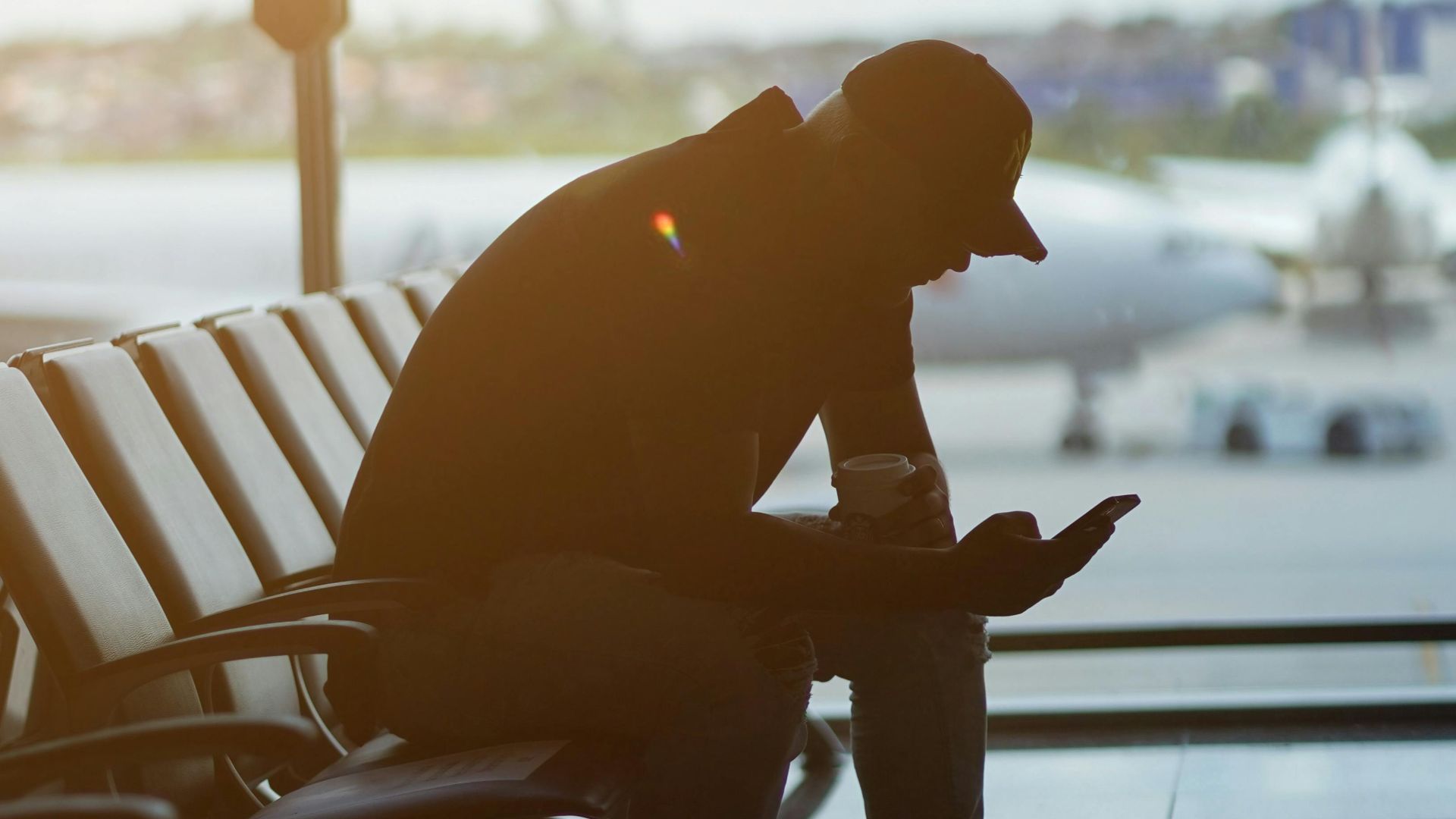Silhouette of a man using a smartphone in an airport waiting area with airplanes visible outside.