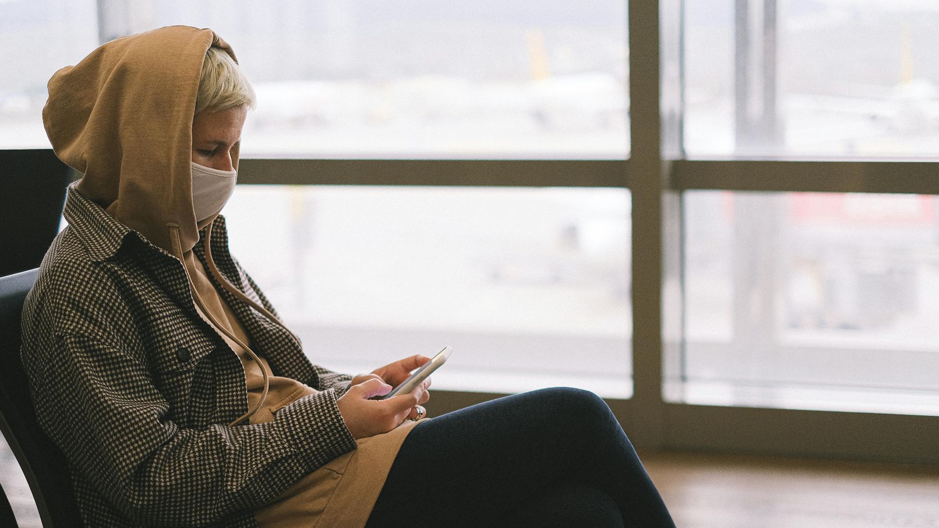 A woman in a hoodie and face mask uses a smartphone while waiting at an airport terminal.