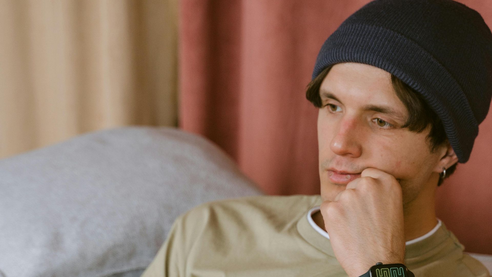 Man wearing a beanie relaxing on a couch while holding a smartphone, looking contemplative.