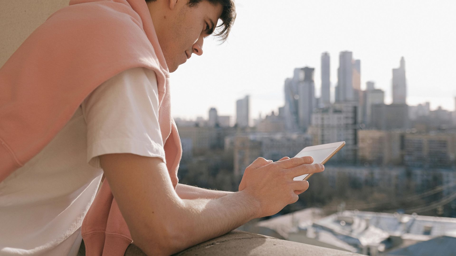A young man stands on a balcony using a tablet with a city skyline in the background.