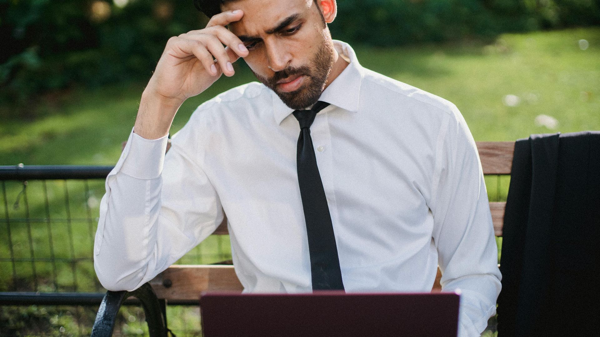 Bearded man in a white shirt and tie working on a laptop on a park bench.